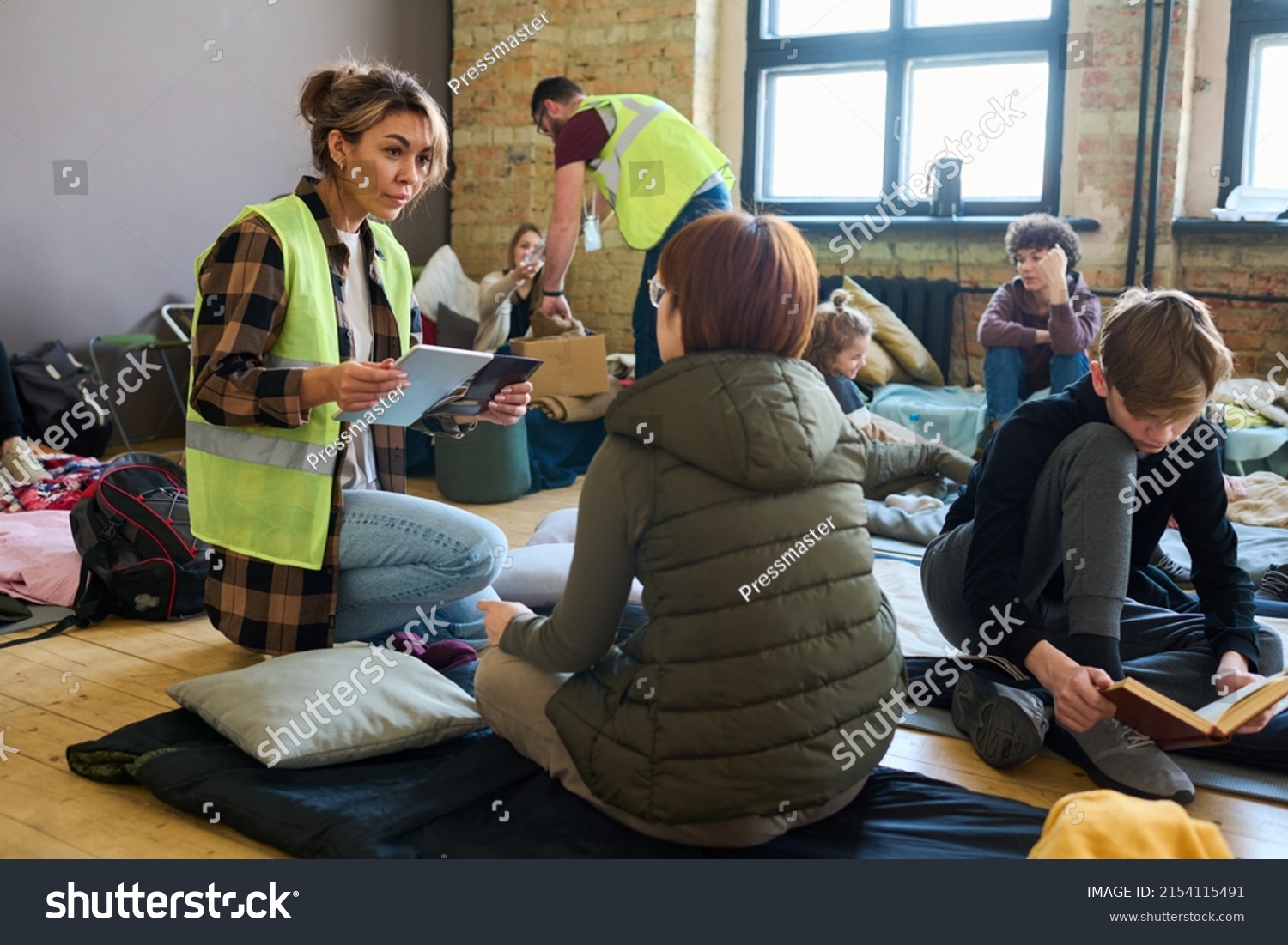 Female volunteer with mobile gadgets communicating to one of refugees while sitting on squats in front of her in shelter for migrants #2154115491