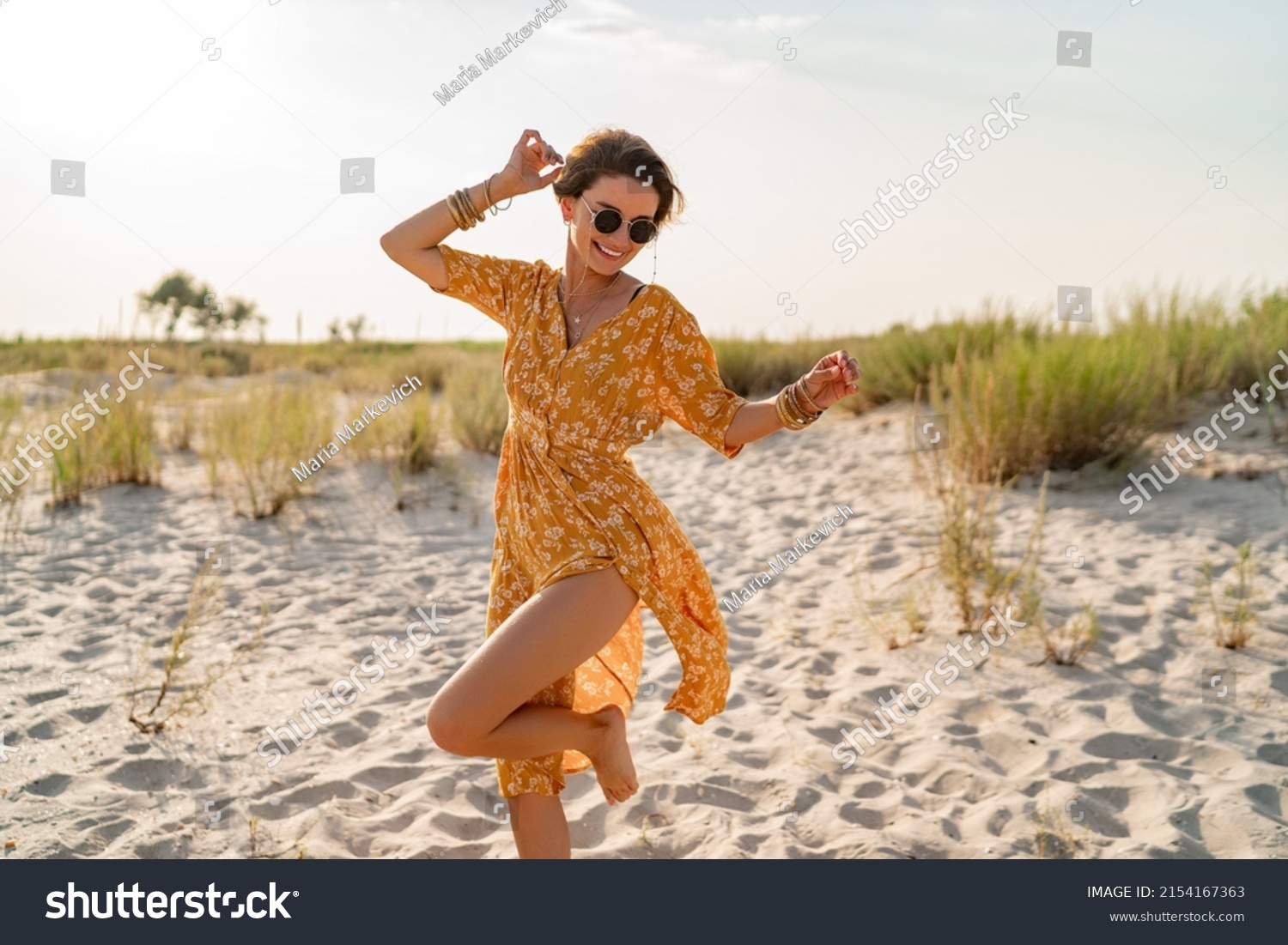 stylish attractive slim smiling woman on beach in summer style fashion trend outfit carefree and happy, feeling freedom, wearing yellow printed dress boho style chic and sunglasses #2154167363