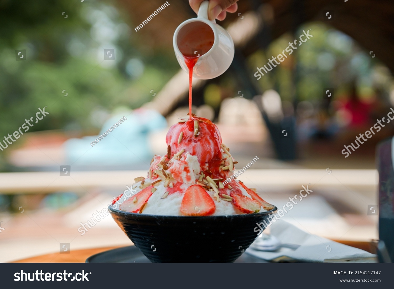 Japanese shaved ice dessert , Hand pouring sweet strawberry sauce on ice cream. Served with strawberry kakigori bingsu topped with almond stick condensed milk. Traditional summer dessert in Japan. #2154217147