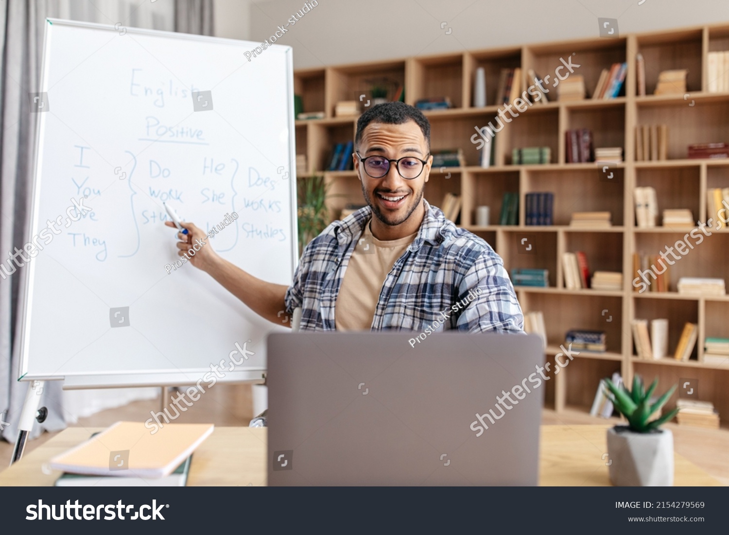 E-learning concept. Excited male English teacher sitting at table, pointing with pen at whiteboard, explaining grammar rules to students, having online lesson from home #2154279569