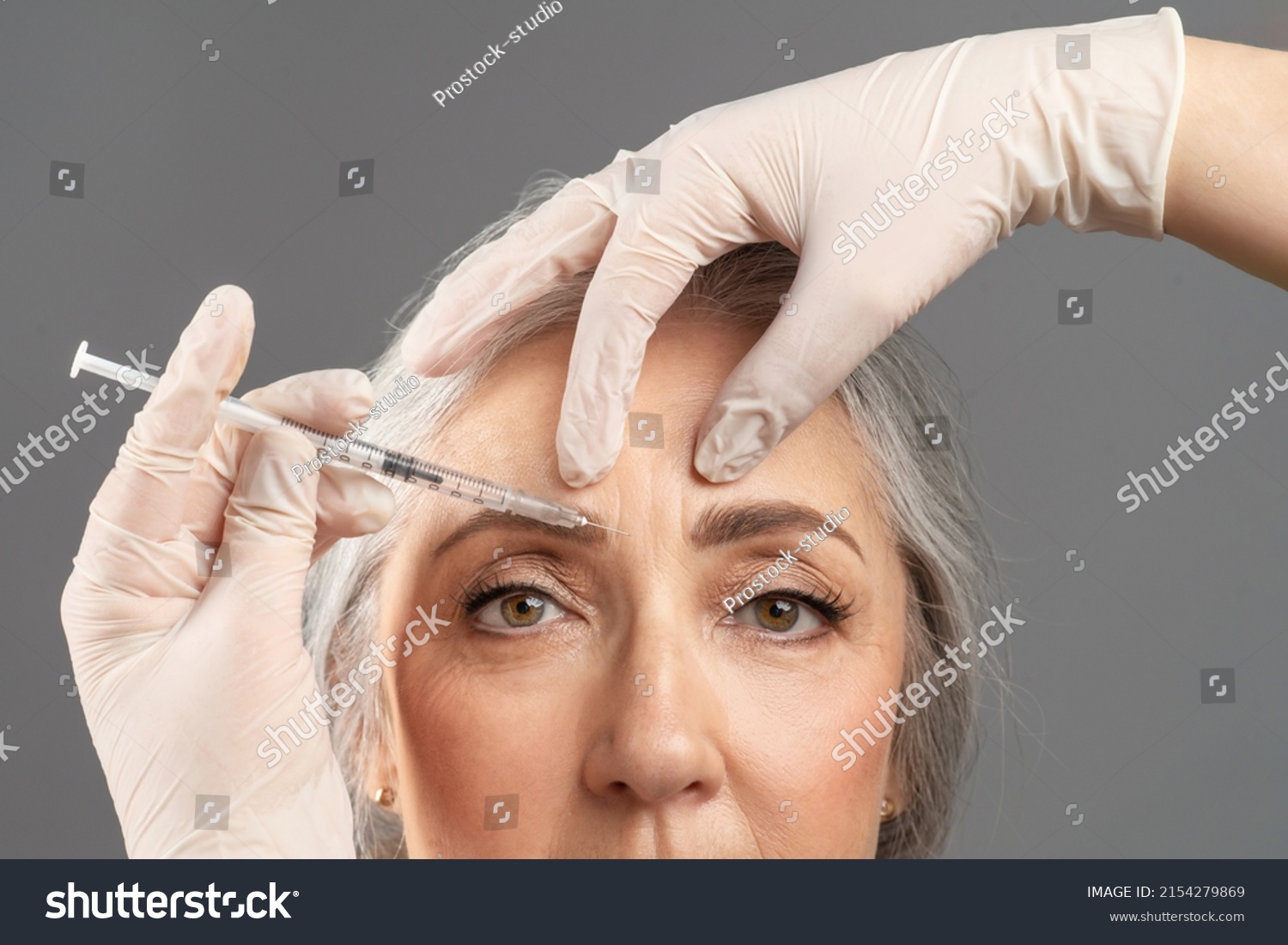 Mature skin rejuvenation. Closeup portrait of senior woman receiving cosmetic treatment with beauty injection on grey studio background. Beautician making anti-aging shot to older lady #2154279869