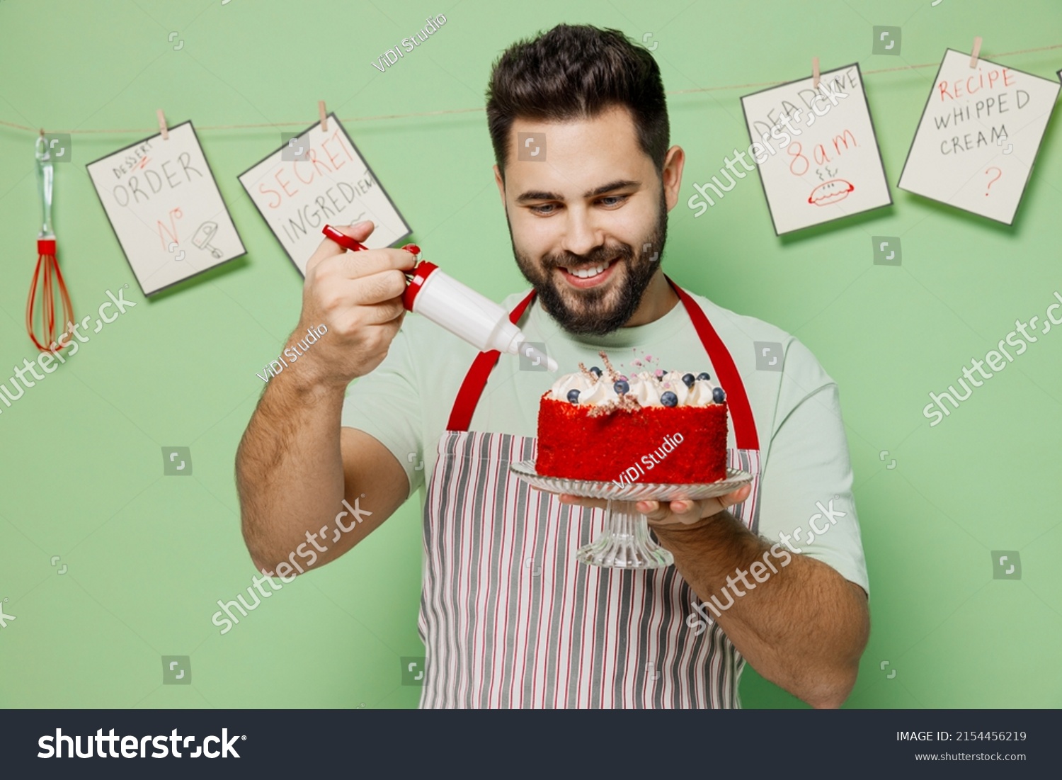Young fun male chef confectioner baker man wear striped apron hold adorn birthday sweet dessert cake with cream isolated on plain pastel light green background studio portrait. Cooking food concept. #2154456219
