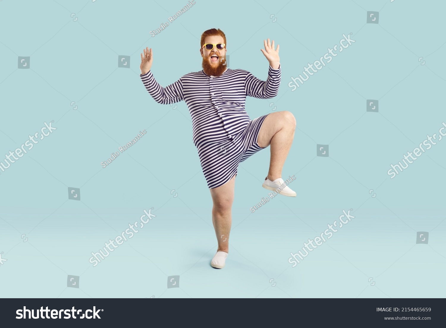 Funny fat guy enjoying his happy summer holiday. Full body length portrait of cheerful goofy chubby bearded man wearing striped retro swimsuit and sunglasses dancing against blue studio background #2154465659