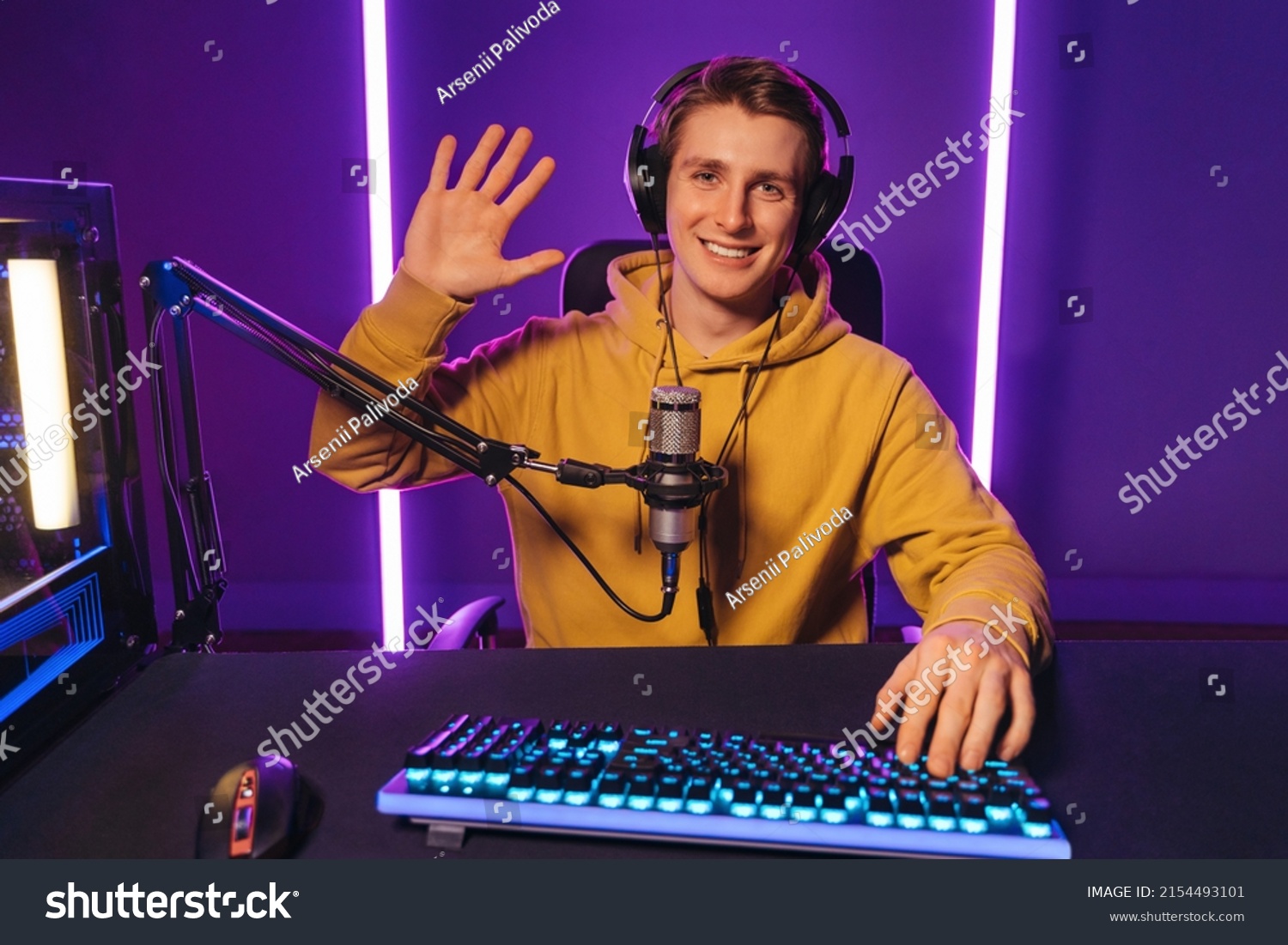 Young handsome vlogger pro gamer waving hand to camera, says hello to his subscribers and followers, smiling on camera. He recording live stream of video game playthrough at home at night. Cyber sport #2154493101