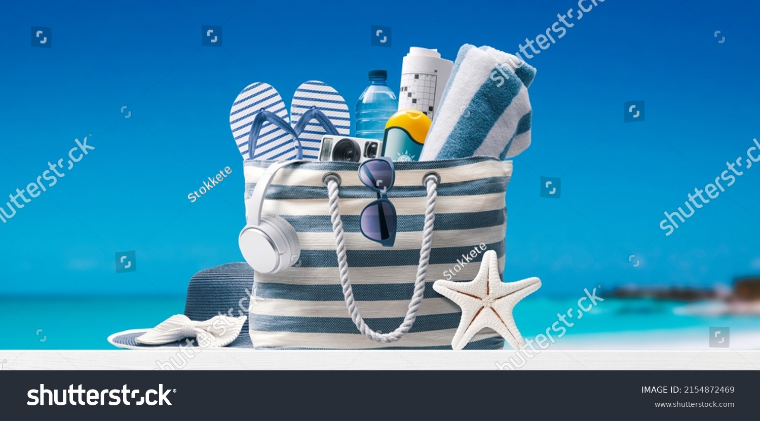 Stylish beach bag with accessories and tropical beach in the background, summer vacations concept #2154872469
