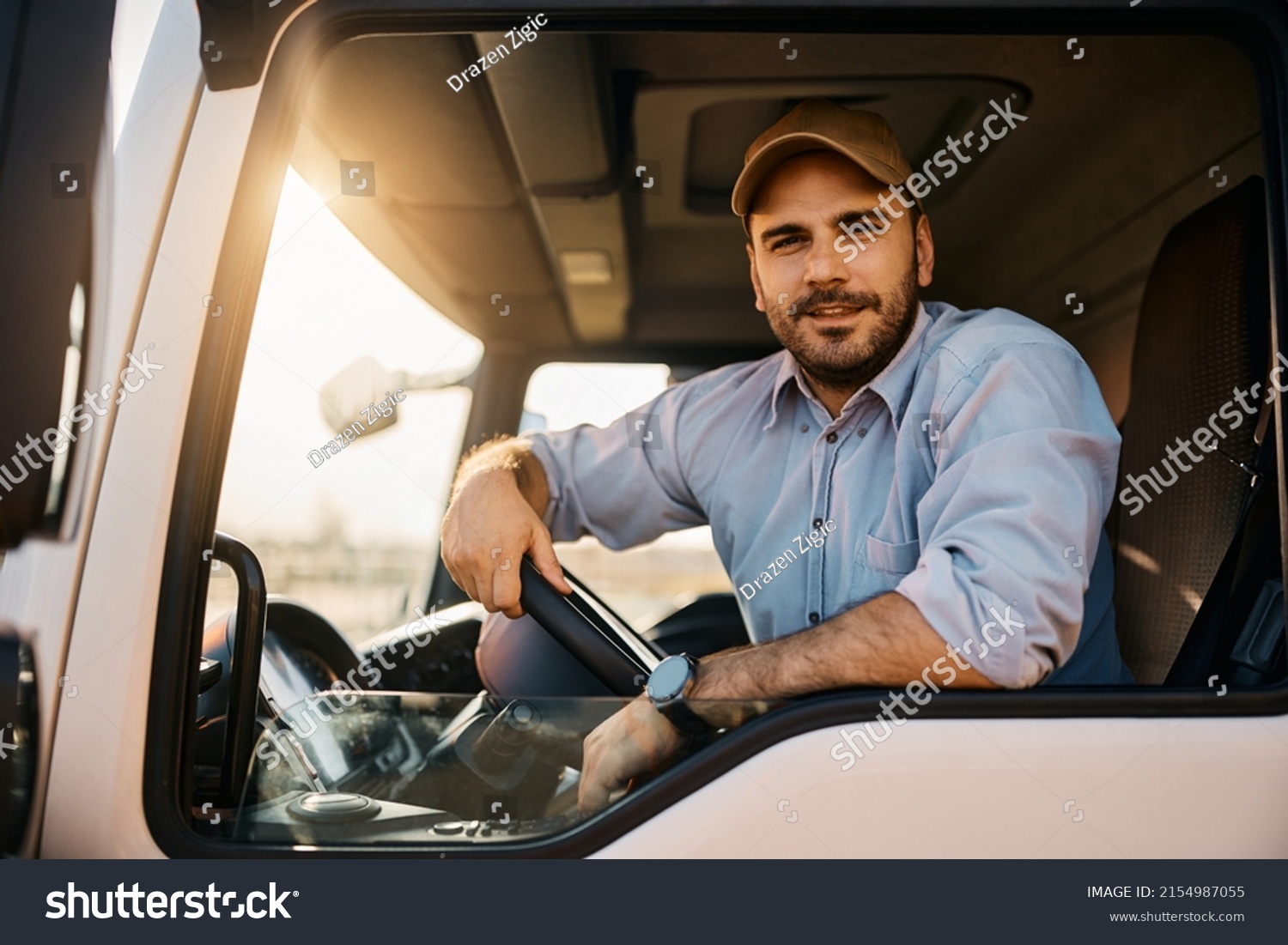 Young truck driver sitting behind steering wheel in a cabin and looking at camera. #2154987055