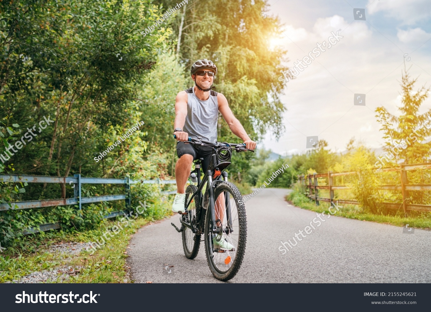 Portrait of a happy smiling man dressed in cycling clothes, helmet and sunglasses riding a bicycle on the asphalt out-of-town bicycle path. Active sporty people concept image. #2155245621