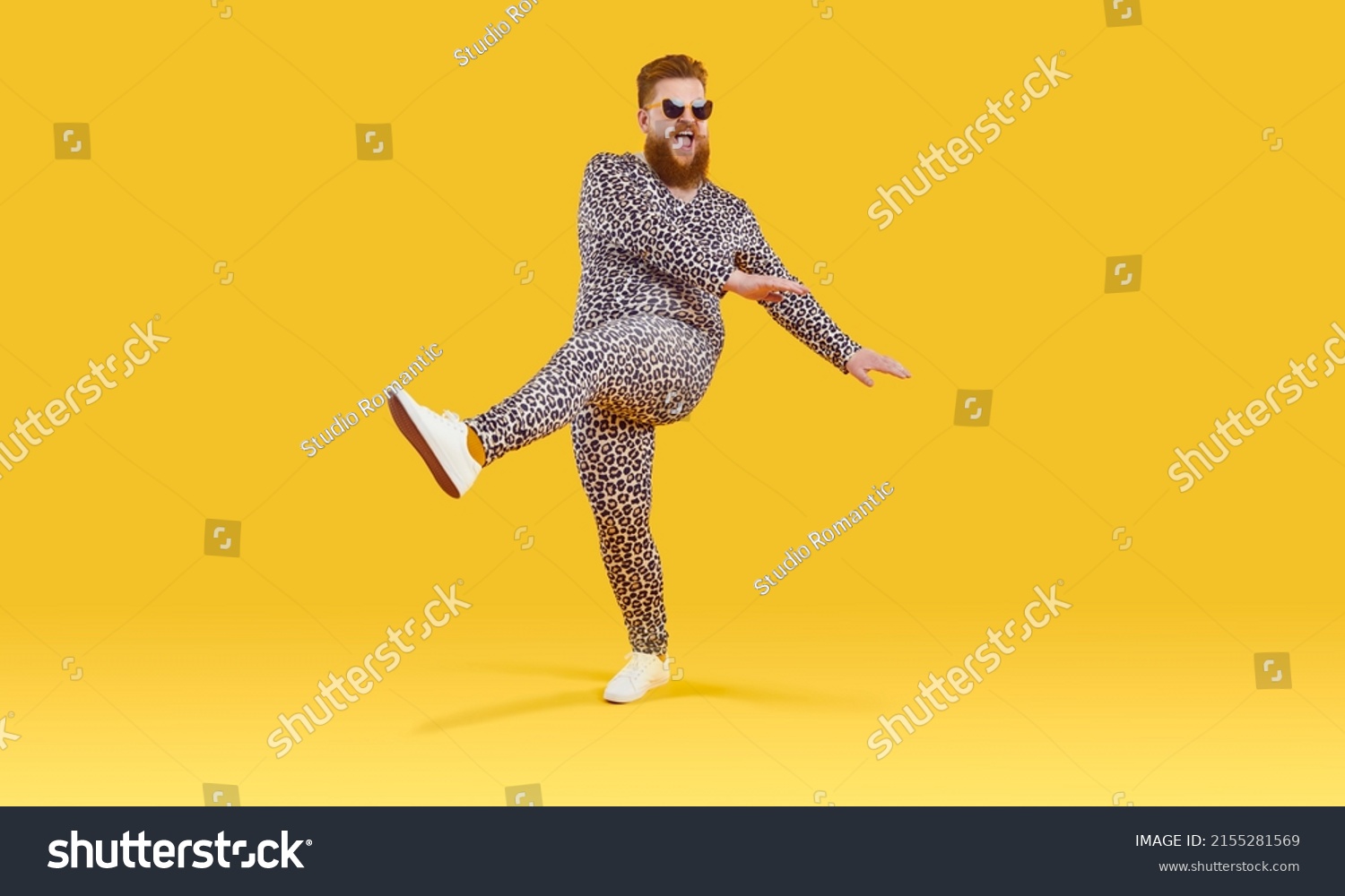 Plus size guy in funny pyjamas having fun in modern studio. Happy carefree fat man wearing comfortable leopard PJs and sunglasses dancing isolated on yellow background. Crazy party and fashion concept #2155281569