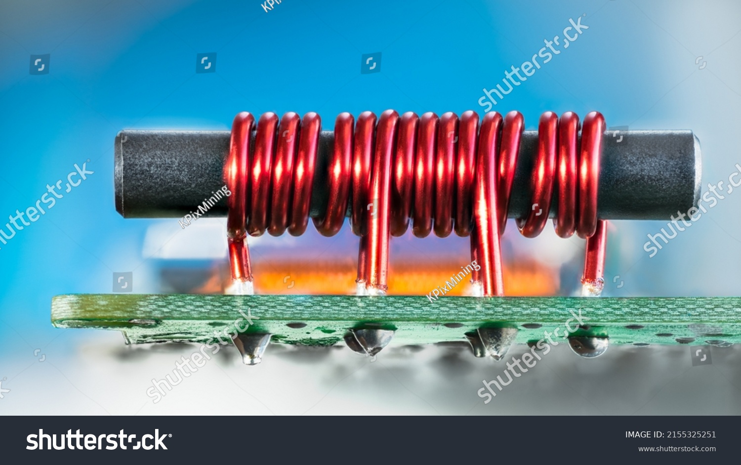 Electronic transformer with ferromagnetic core and coil copper wire on PCB. Closeup of red cylindric inductor soldered on green circuit board with tin on bottom side and blue background. Cable TV amp. #2155325251