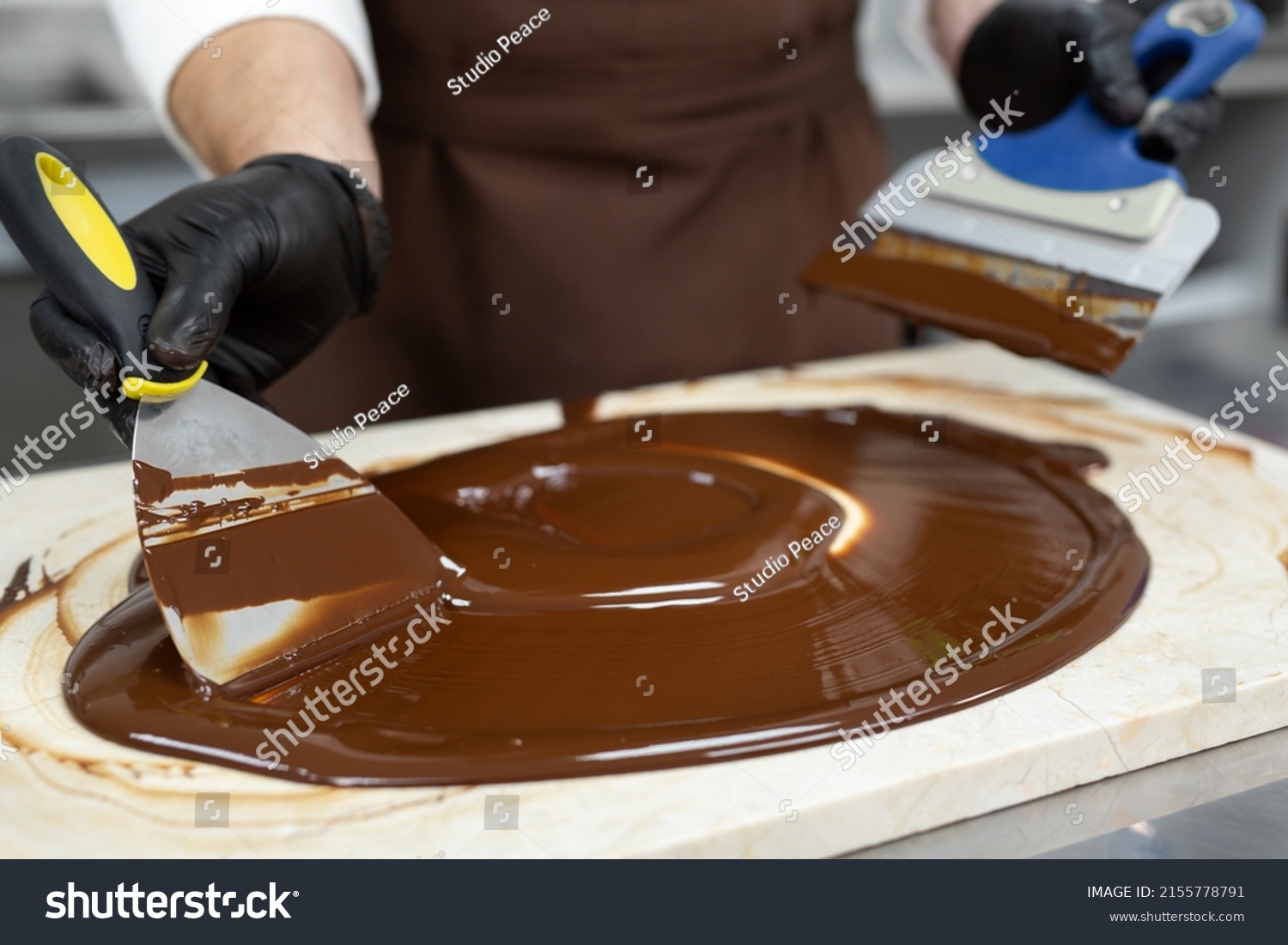 Male chocolatier uses a spatula to stir the tempered liquid chocolate on a granite table #2155778791
