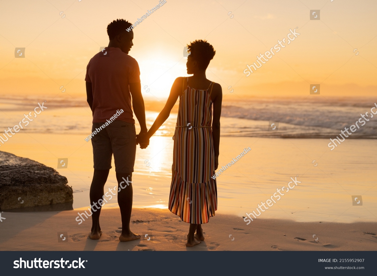 Rear view of african american young couple holding hands and standing on beach against sky at sunset. nature, unaltered, beach, love, togetherness, lifestyle, enjoyment and holiday concept. #2155952907