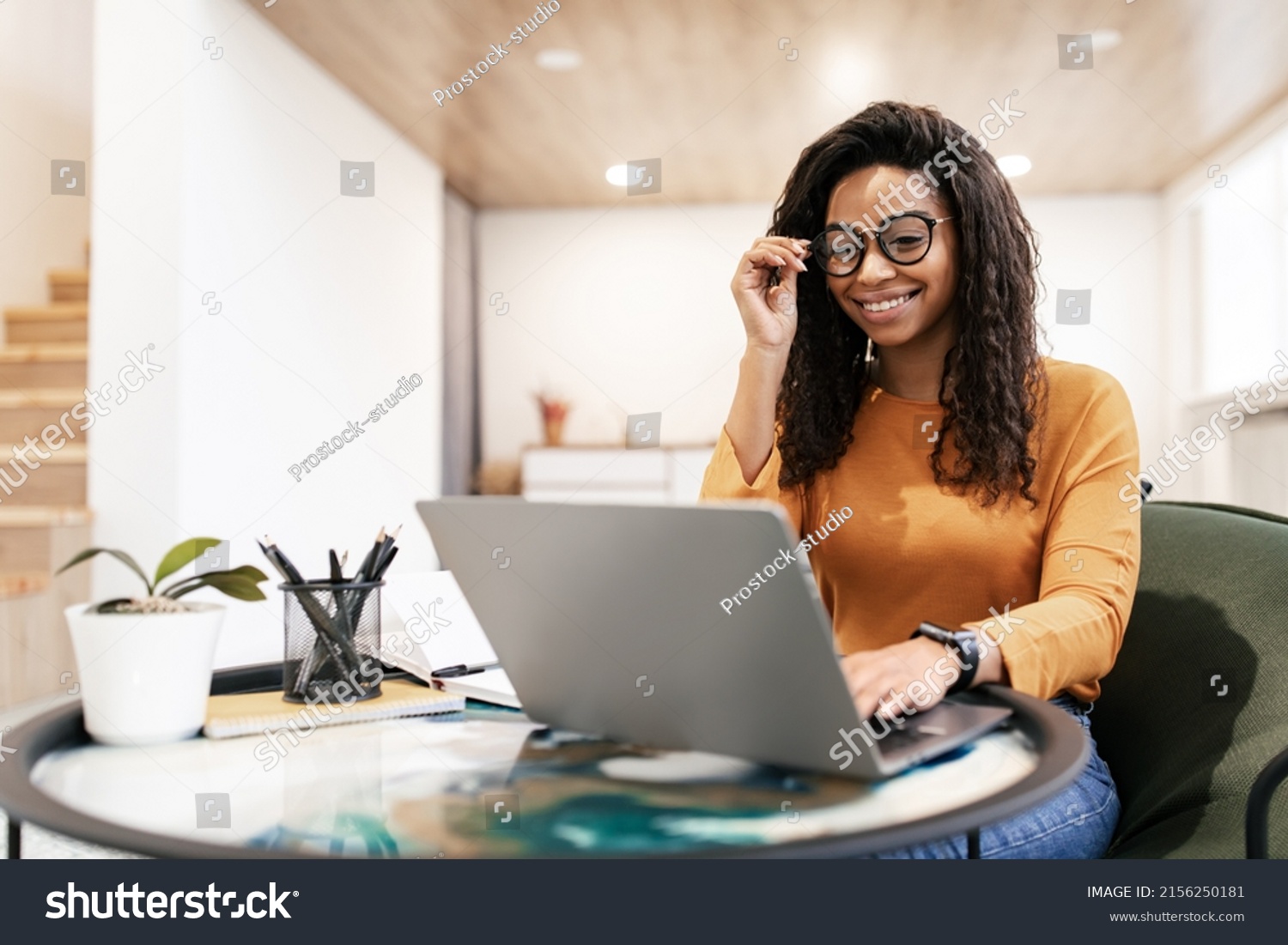People And Technology. Portrait of young black woman wearing eyeglasses using pc sitting at table on beanbag chair in living room, typing on keyboard. Cheerful lady browsing internet, free copy space #2156250181