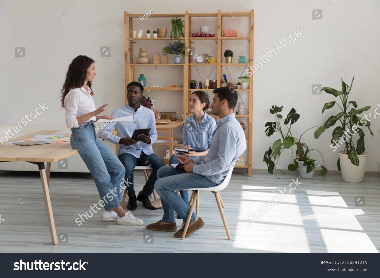 Diverse millennial business team meeting in modern office eco interior, sitting in circle, talking, holding documents, gadgets. Boss discussing project with employees. Mentor training interns #2156291215