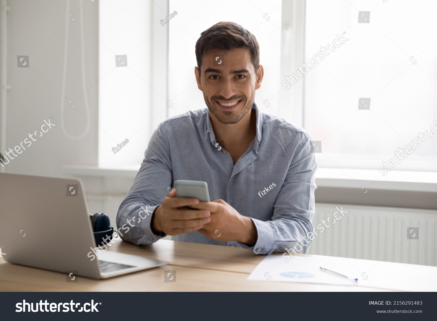 Happy digital gadgets user man using smartphone, laptop at home workplace head shot portrait. Young handsome business man holding mobile phone, sitting at computer, looking at camera #2156291483