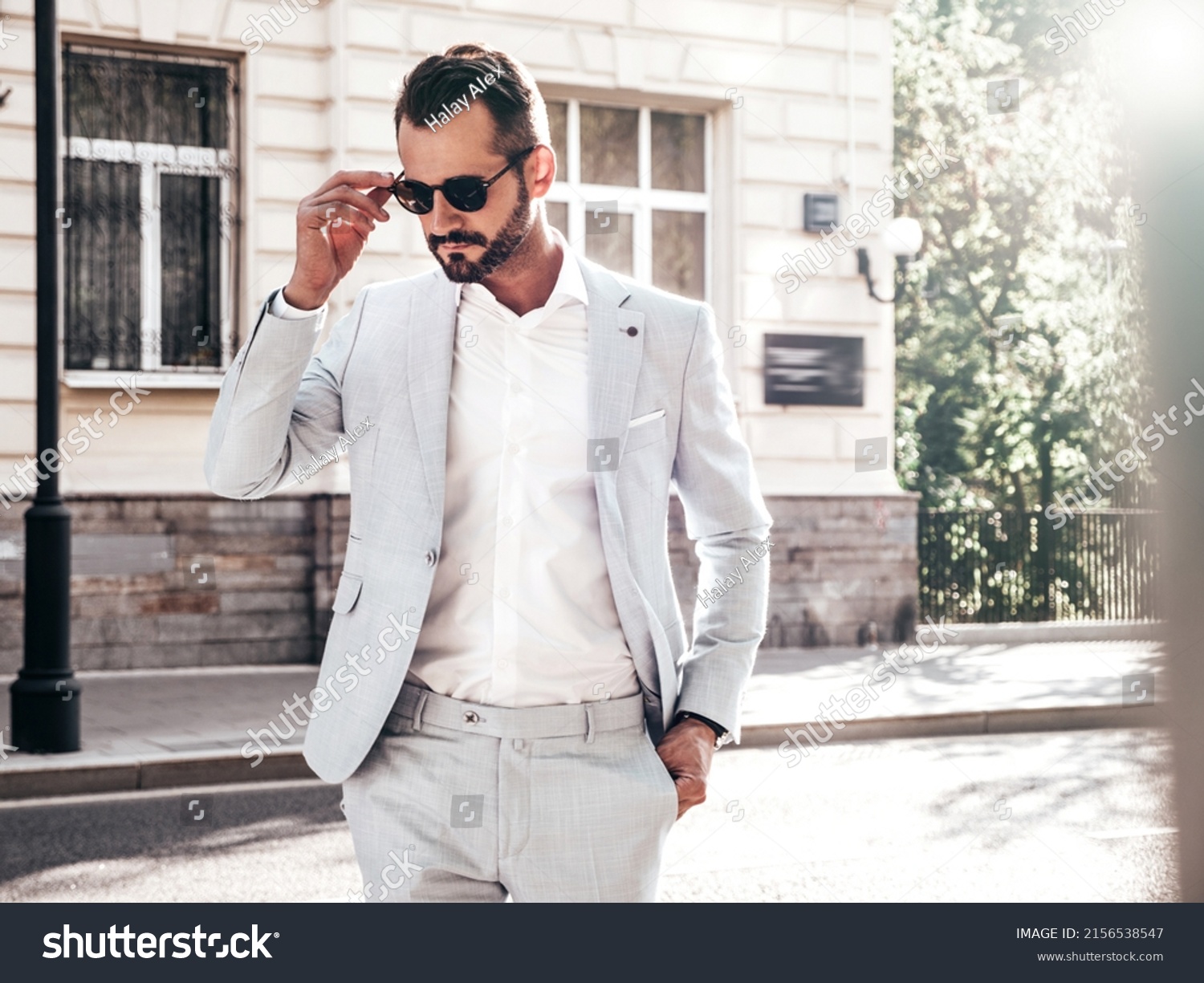 Portrait of handsome confident stylish hipster lambersexual model.Sexy modern man dressed in elegant white suit. Fashion male posing in the street background in Europe city at sunset. In sunglasses #2156538547