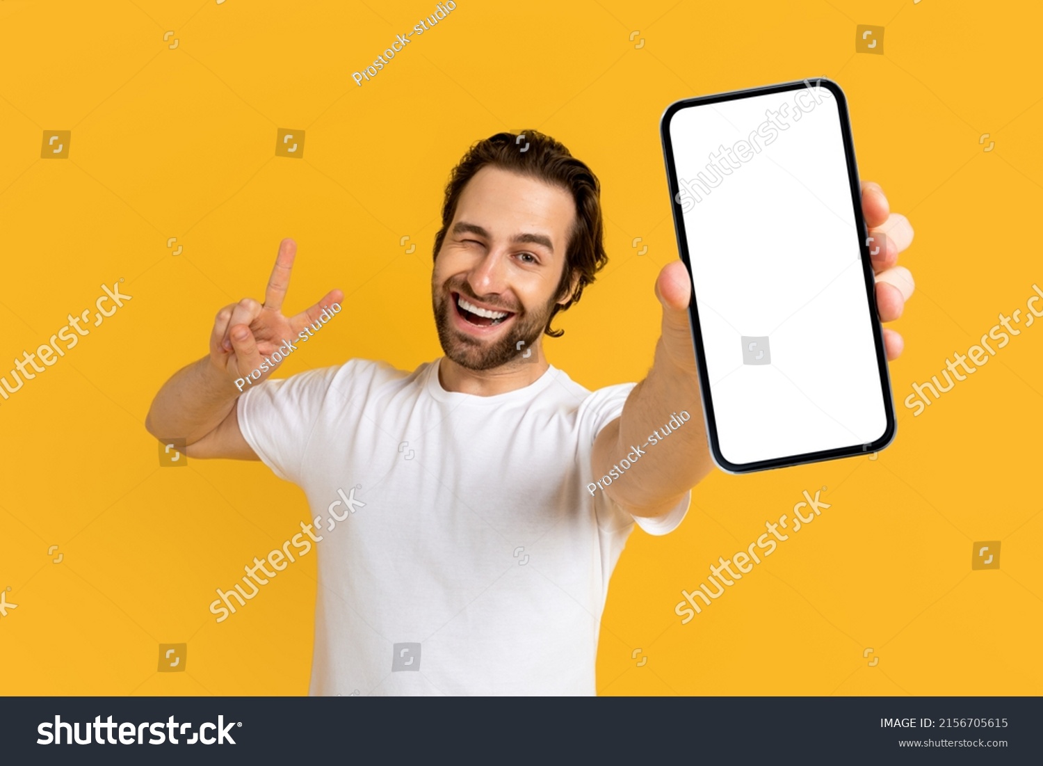 Cheerful emotional young european guy in white t-shirt show phone with empty screen and peace sign with hand isolated on yellow background, studio. Gesture, big sale, new app, device, app and offer #2156705615