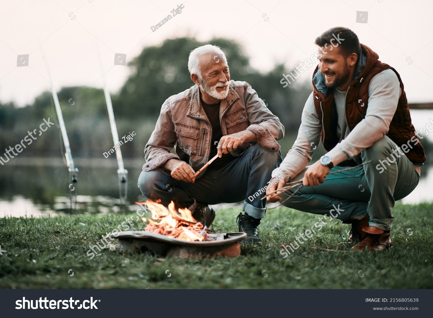 Happy mature man and his adult son talking while relaxing by campfire during their fishing day. #2156805639