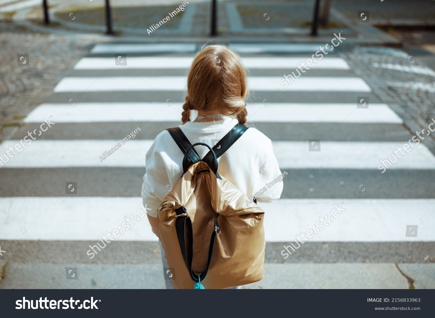 Seen from behind trendy pupil in white sweatshirt with backpack crossing crosswalk and going to school outdoors in the city. #2156833963