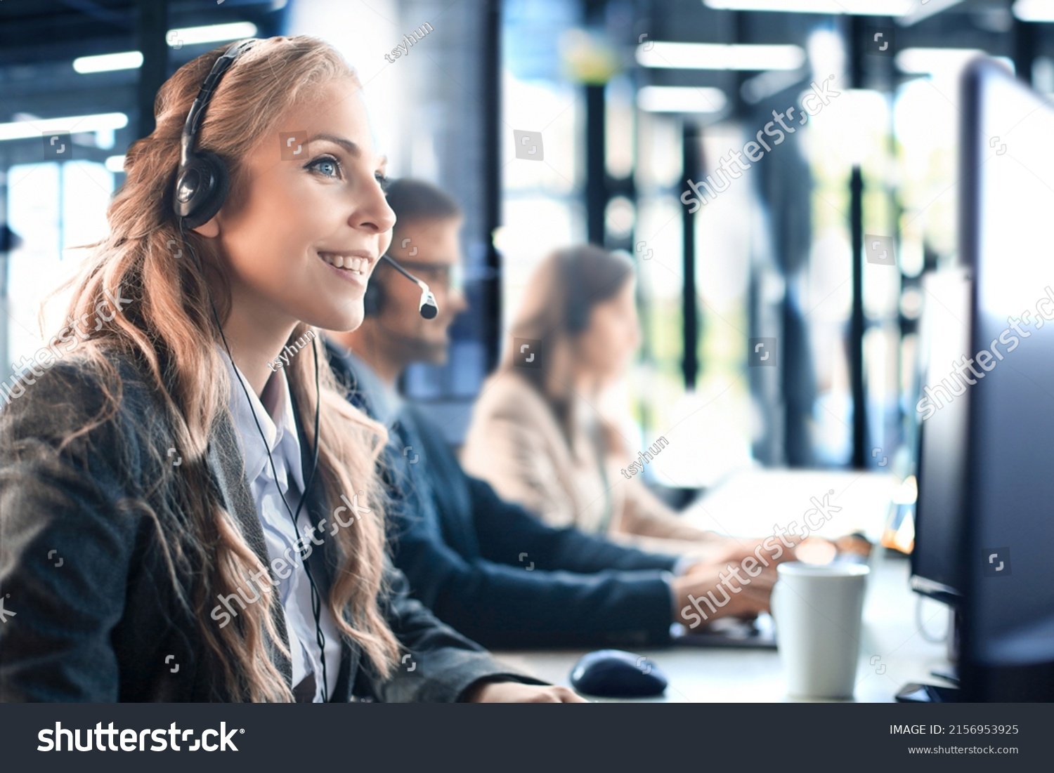 Female customer support operator with headset and smiling, with collegues at background. #2156953925