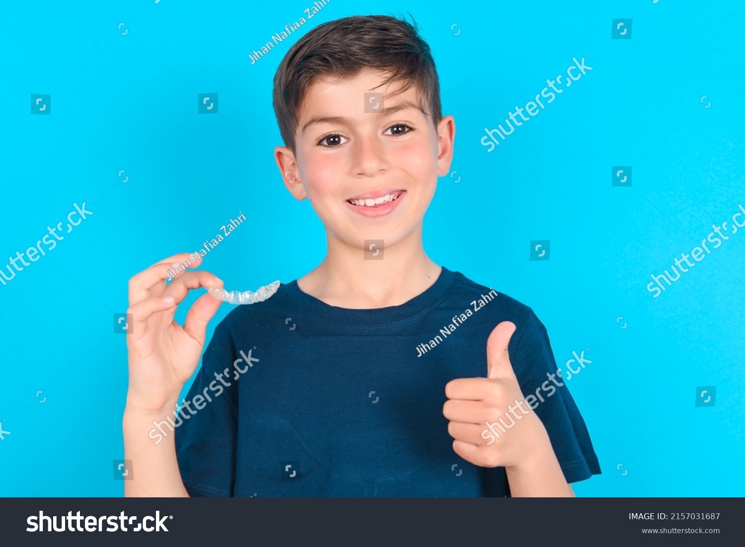 caucasian kid boy wearing blue T-shirt over blue background holding an invisible braces aligner and rising thumb up, recommending this new treatment. Dental healthcare concept. #2157031687