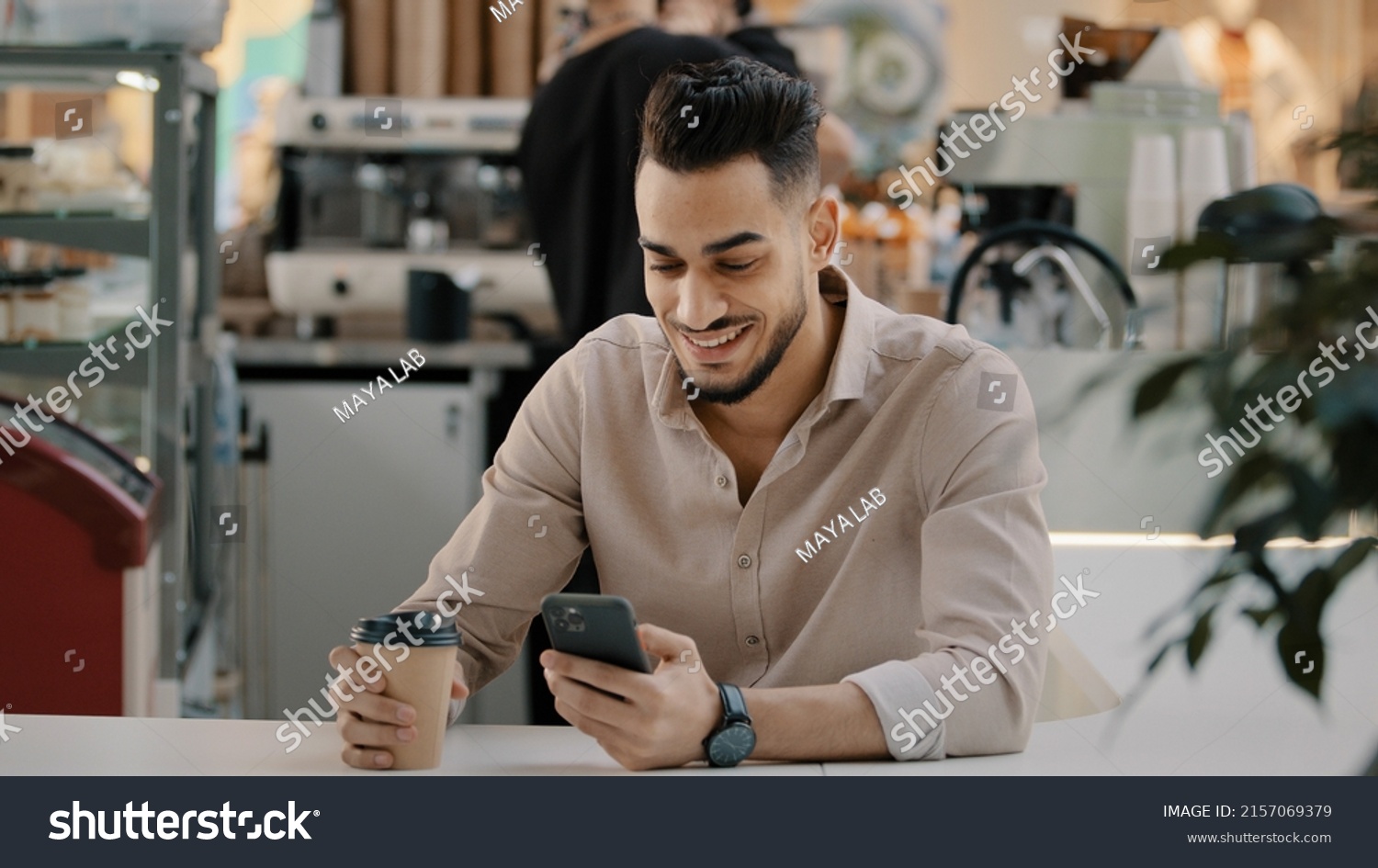 Indian hispanic arabian man multiracial businessman sitting at cafe table drinking hot coffee tea from paper cup looking in mobile phone typing smartphone chatting browsing using online app free wi-fi #2157069379