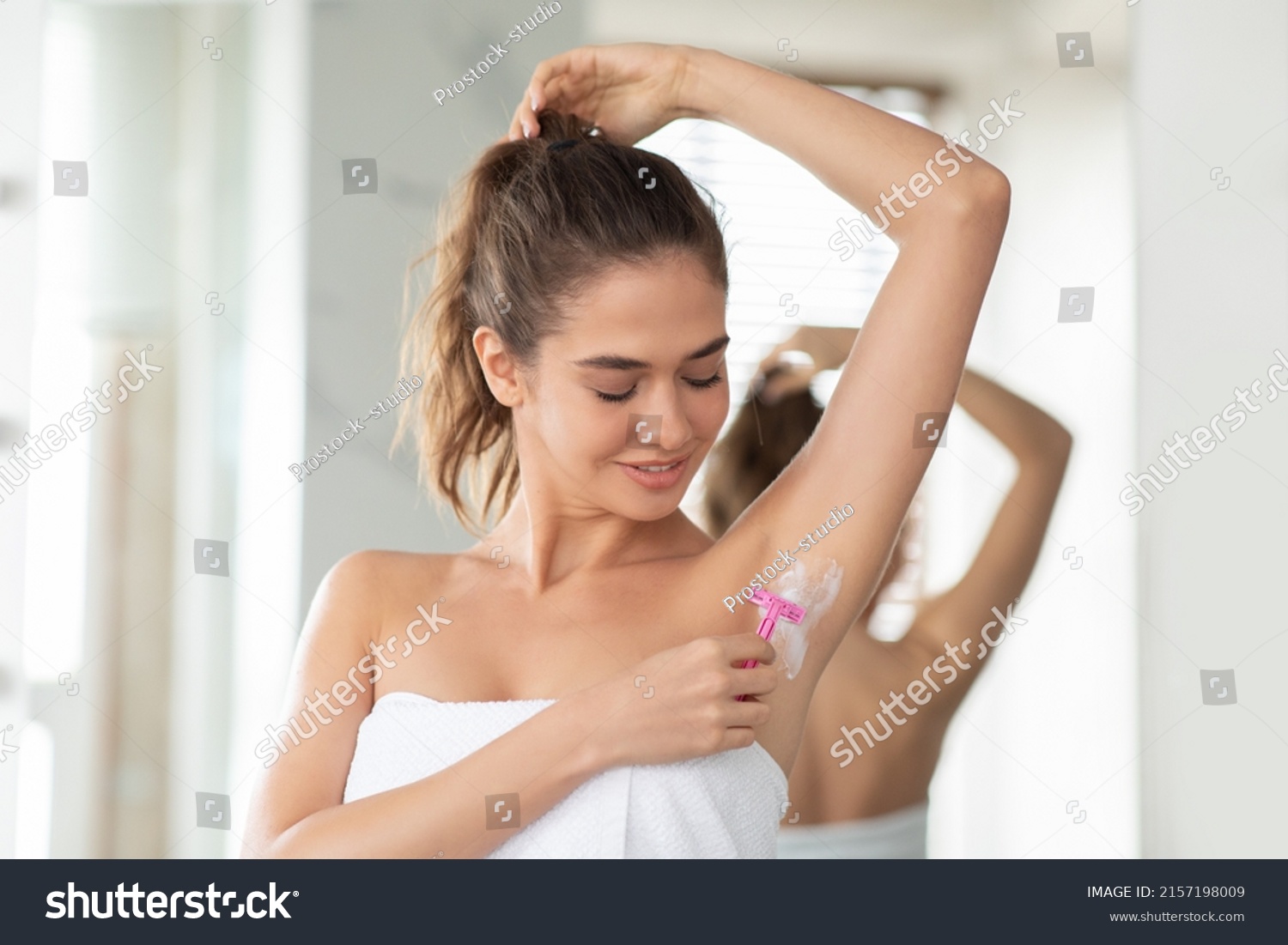 Young Lady Shaving Armpits Covered With Foam Removing Hair Standing Wrapped In Towel In Modern Bathroom. Woman Raising Arm Making Underarms Depilation. Hair Removal Concept #2157198009