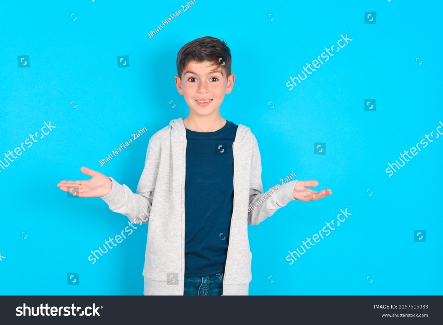 So what? Portrait of arrogant caucasian kid boy wearing grey hoodie over blue background shrugging hands sideways smiling gasping indifferent, telling something obvious. #2157515983