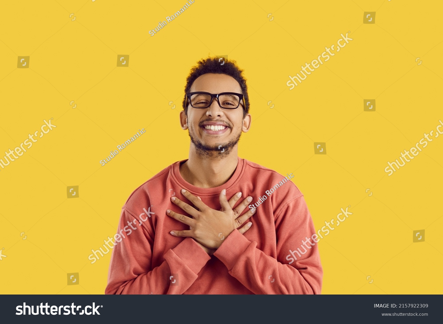 Happy black man in glasses smiling and holding his hands on his chest to express endless gratitude and appreciation. Cheerful ethnic guy says thank you from the bottom of his heart for gift or present #2157922309