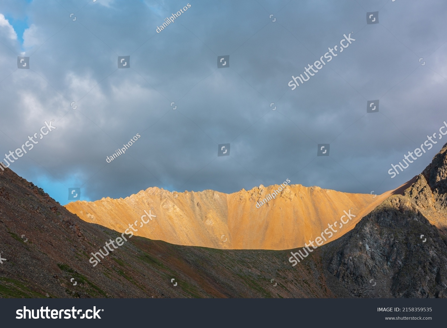 Dramatic alpine landscape with sunlit wide sharp mountain ridge under overcast sky at changeable weather. Atmospheric mountain scenery with large sharp rocks on ridge top in sunlight under cloudy sky. #2158359535