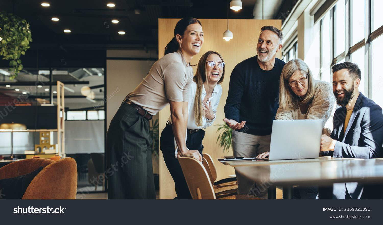 Happy businesspeople laughing while collaborating on a new project in an office. Group of diverse businesspeople using a laptop while working together in a modern workspace. #2159023891