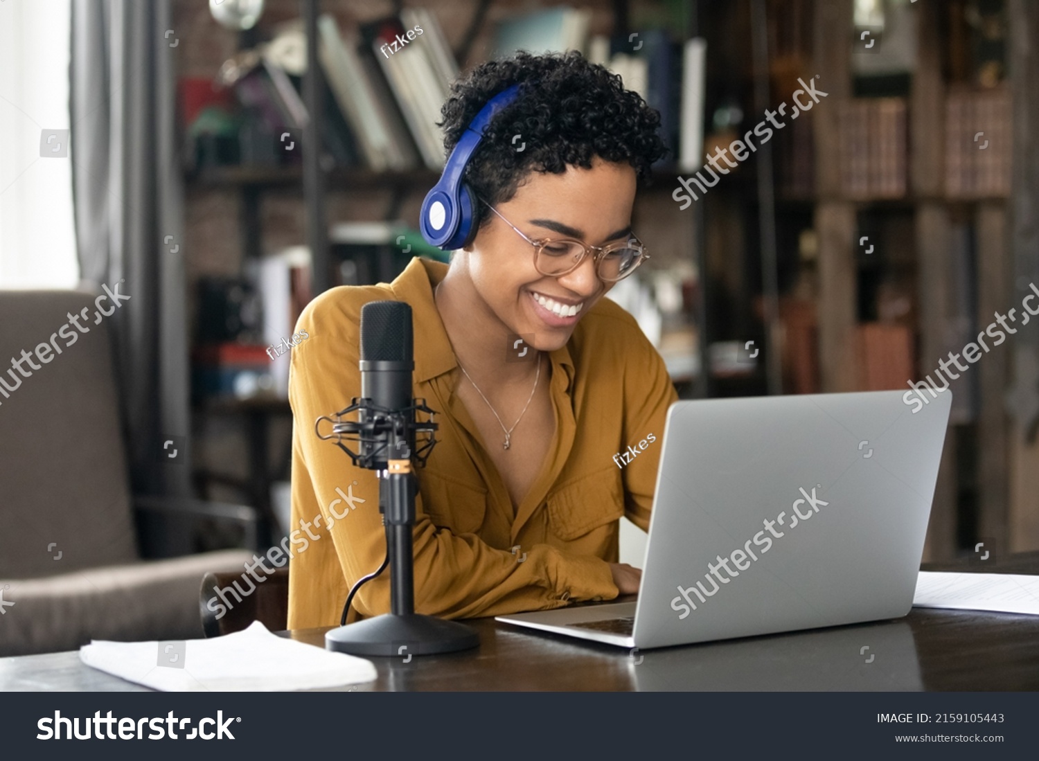 Smiling young African woman podcaster in headphones sit at desk in front of laptop and microphone. Share information, make audio podcast for internet audience, lead on-line stream, blogging concept #2159105443