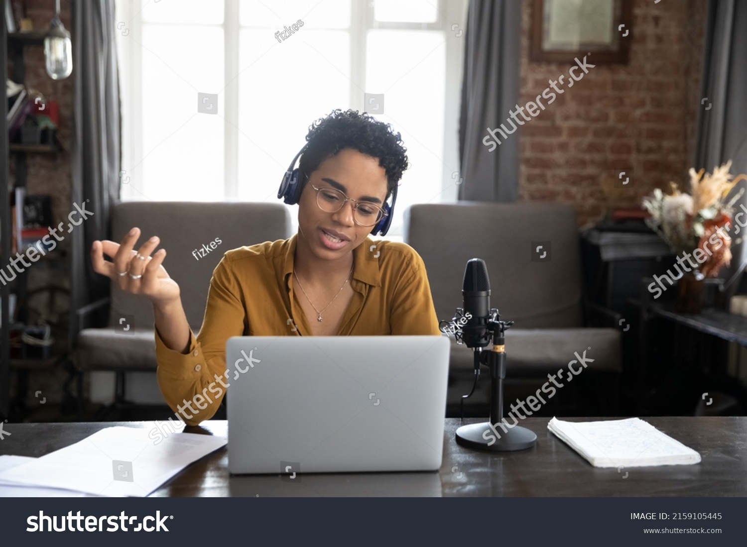 African woman wear wireless headphones sit at desk looks at laptop screen make speech talk into mike, records or lead live stream via internet in real time. Podcast, online radio program event concept #2159105445