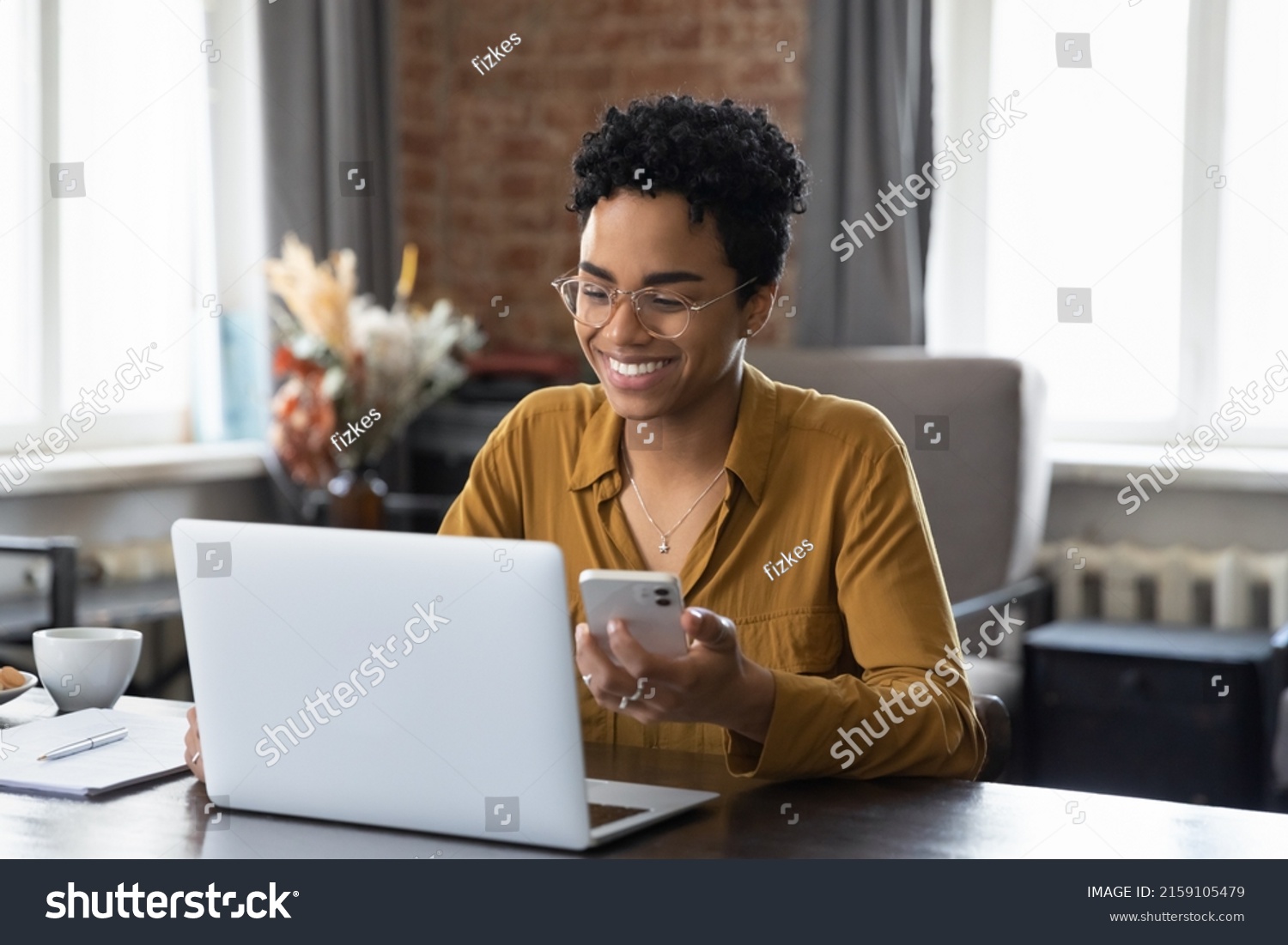 African woman sit at workplace desk holds cellphone staring at laptop, synchronize data between computer and gadget in office, use corporate devices and business application, plan work, use organizer #2159105479