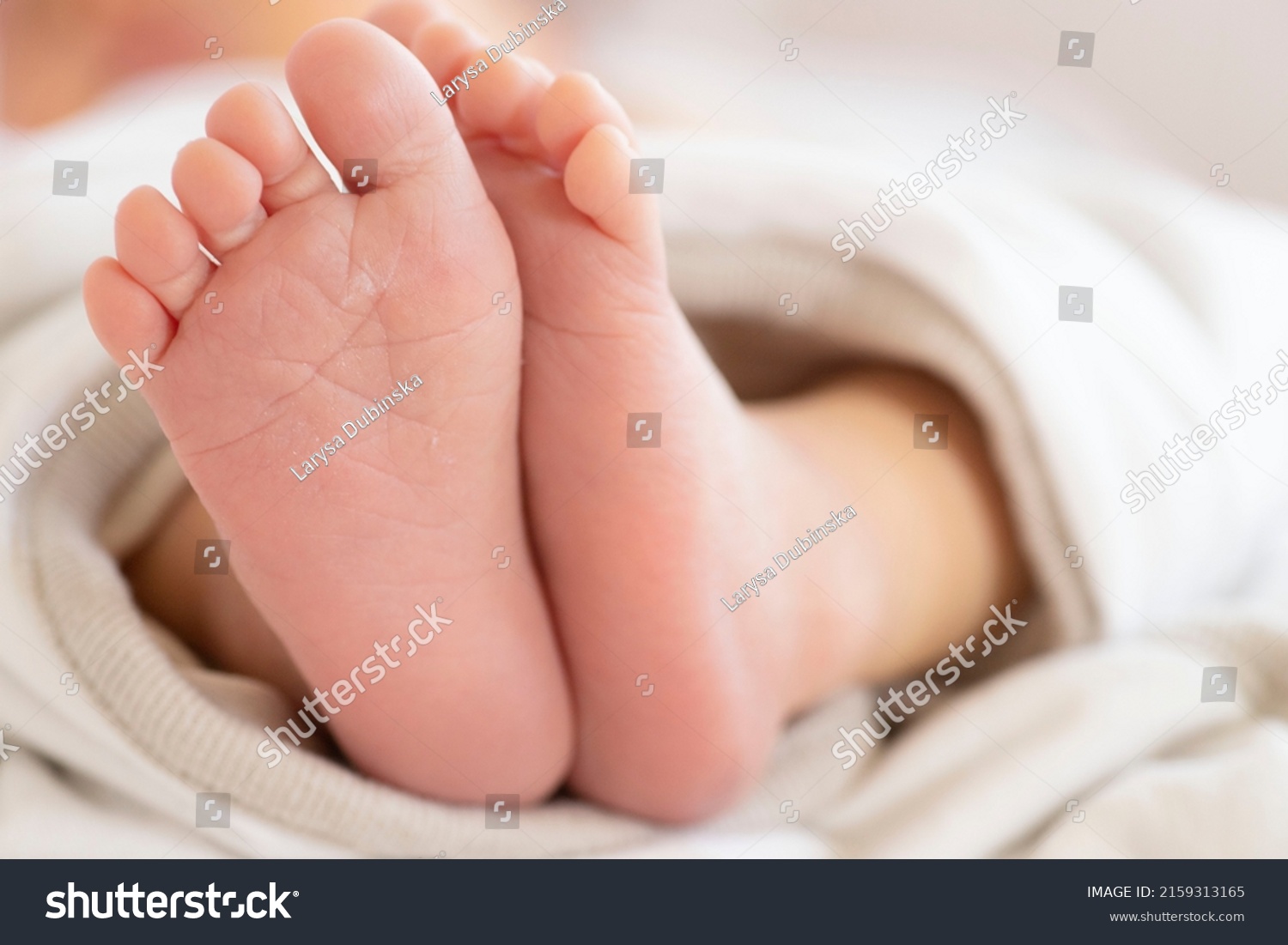 Small bare feet of a newborn covered with a cotton blanket. Daylight. Lifestyle. The concept of motherhood, childhood, parenthood, the first months of a baby's life, the delicate skin of a baby. #2159313165
