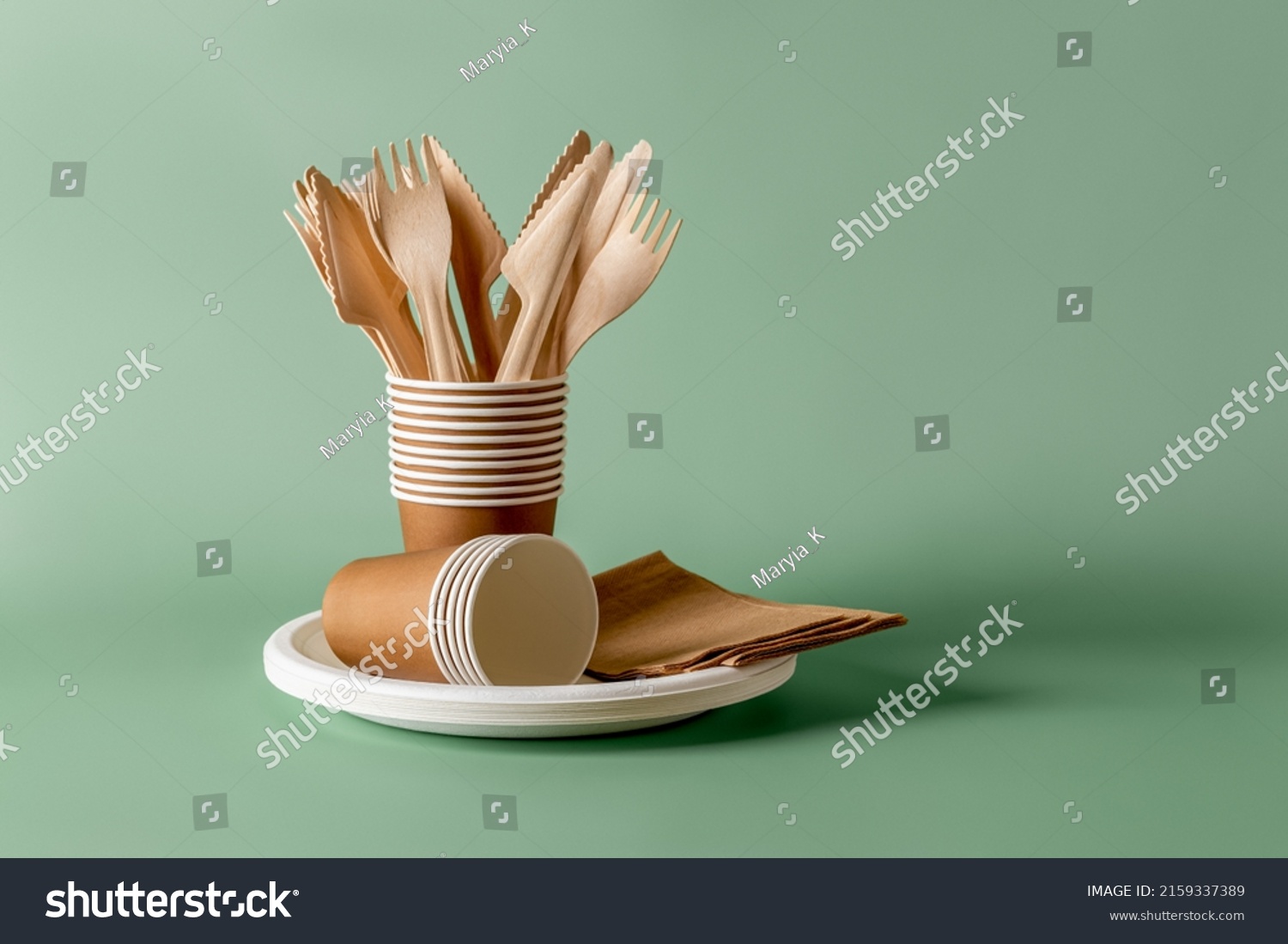 Stack of eco-friendly disposable tableware. Wooden forks and knives, paper cups and plates against green background. Biodegradable cutlery and dishes for picnics, takeaways. Copy space. Front view. #2159337389
