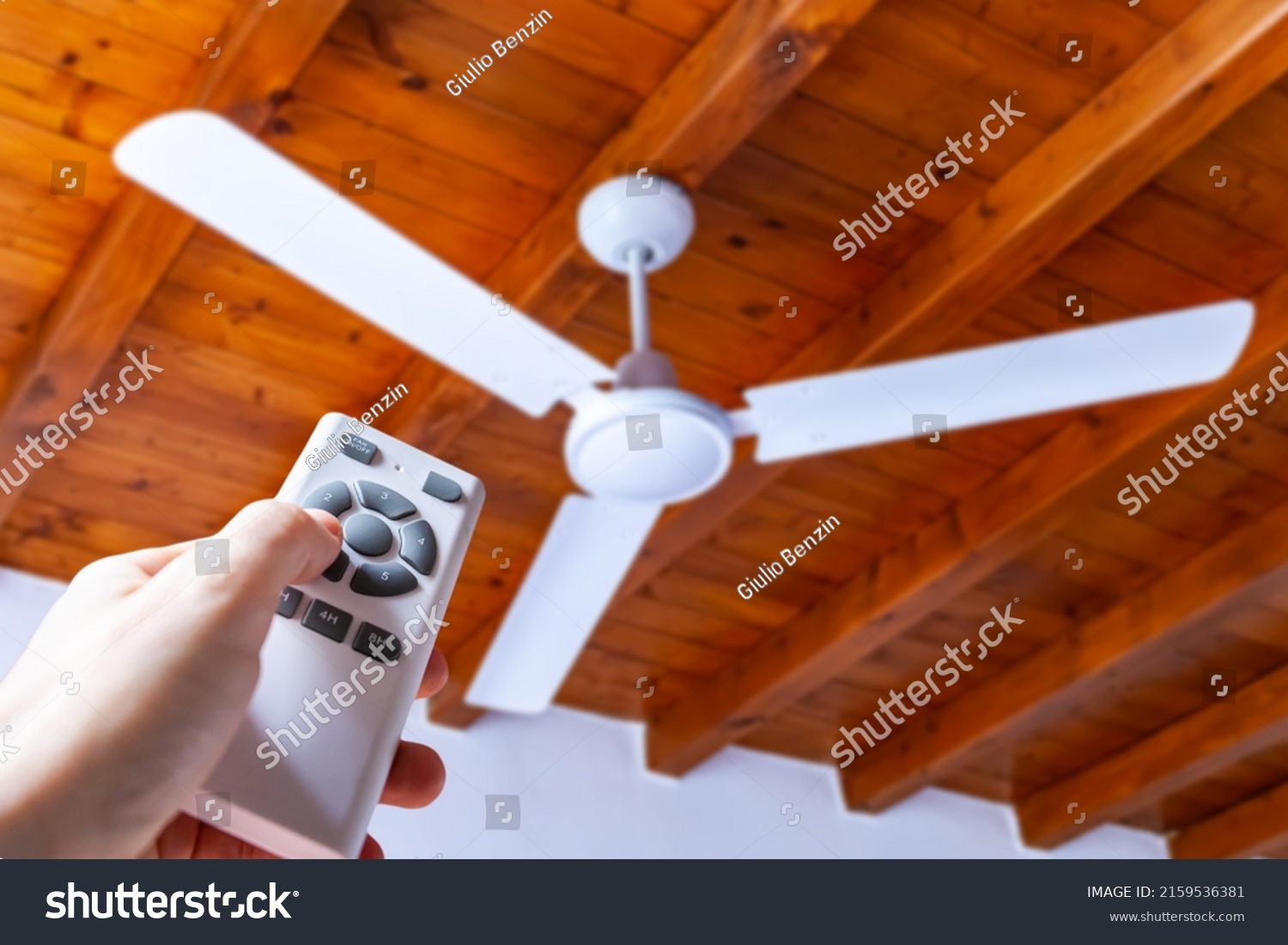 A man uses a remote control to turn on a white ceiling fan mounted in a house with wooden ceilings.	 #2159536381