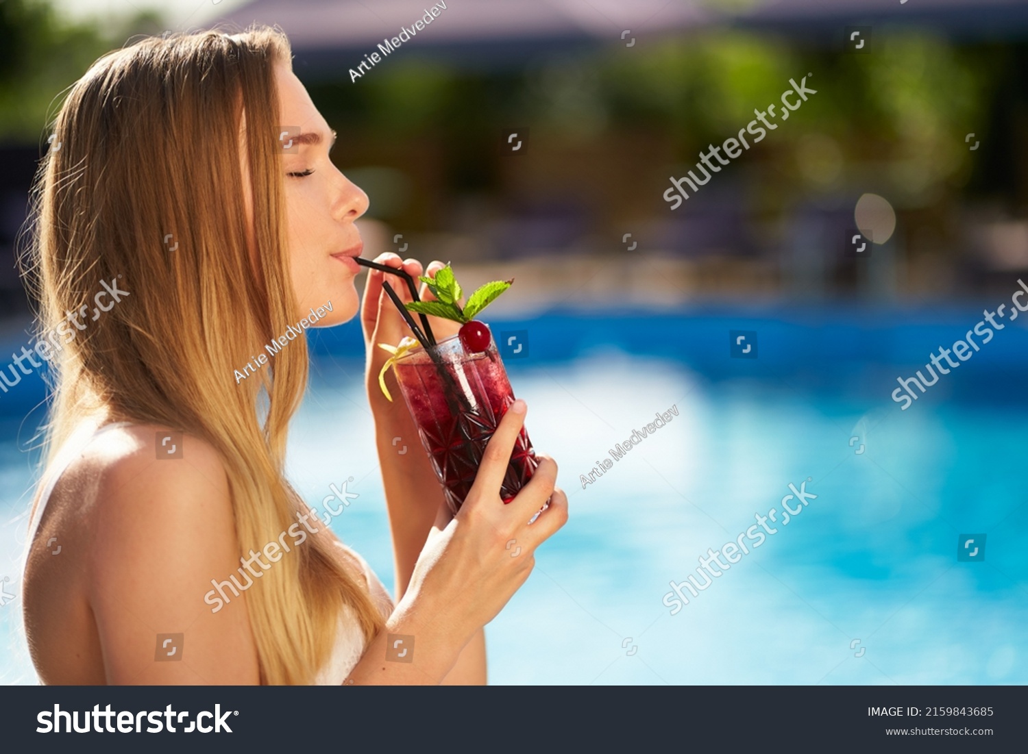 Pretty woman drinks refreshing cocktail decorated with mint, cherry and lemon. Female enjoys a drink near swimming pool with blue water. Girl chilling with beverage in tropical sun. Vacation concept #2159843685