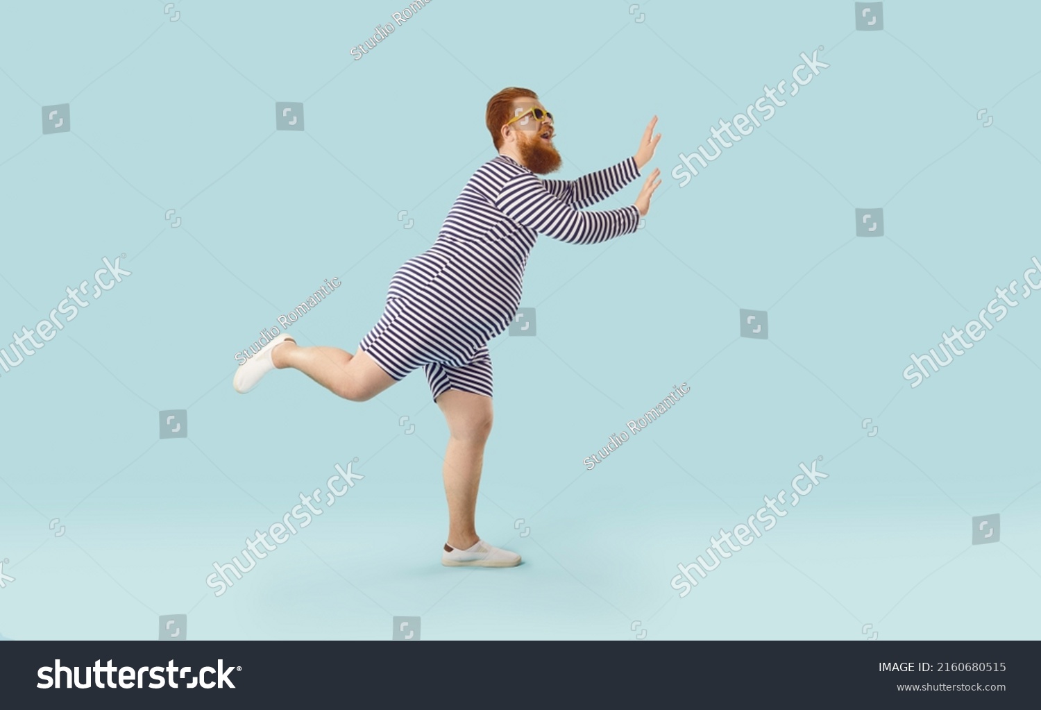 Profile view of funny fat ginger man wearing striped swimsuit and sunglasses dancing and having fun isolated on light blue colour background. Summer holiday, vacation, beach party concept #2160680515