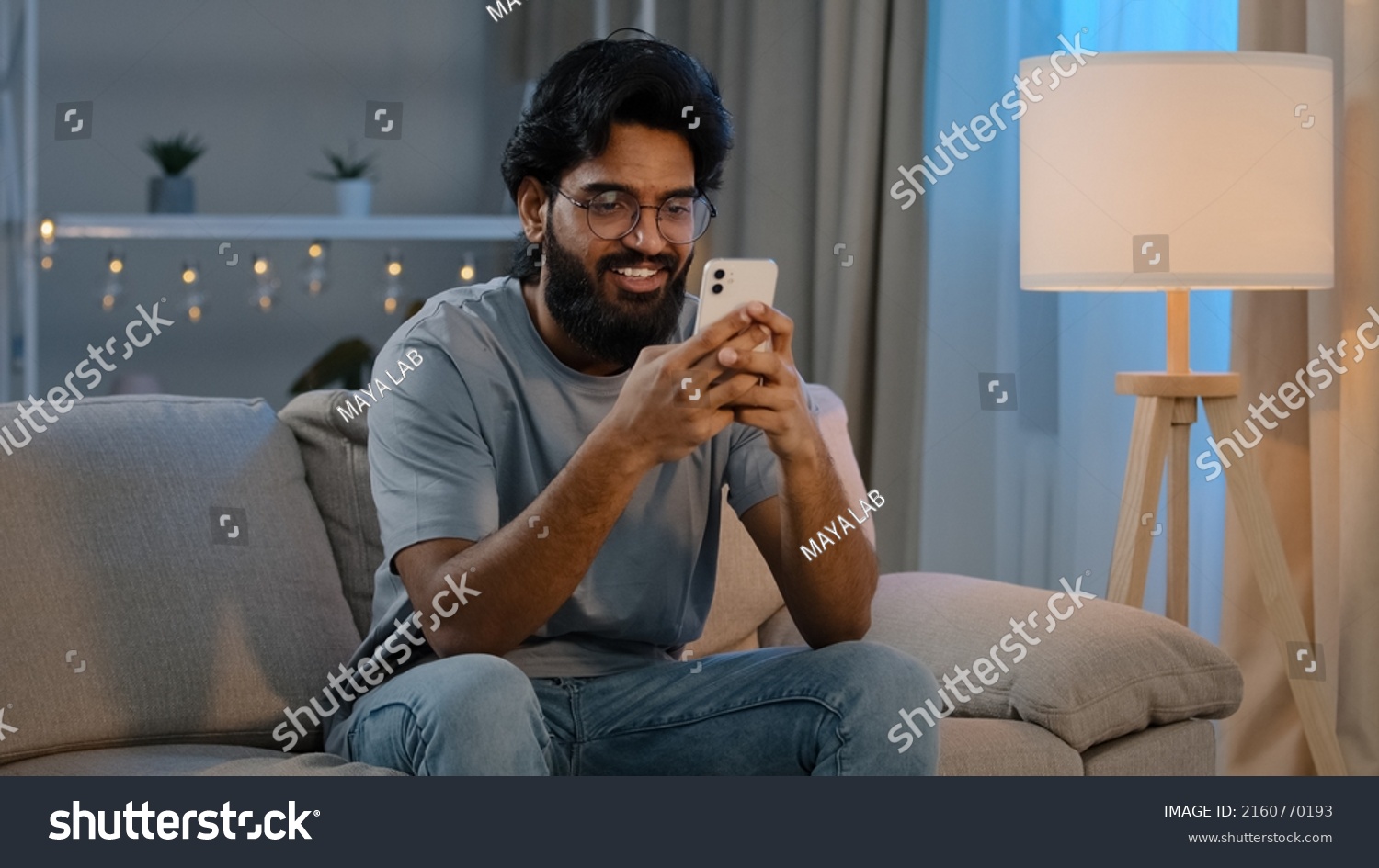 Smiling Arabian Indian man bearded male looking in smartphone smile texting with friend girlfriend messaging chatting at home sitting on couch at night playing game in mobile app watching funny video #2160770193