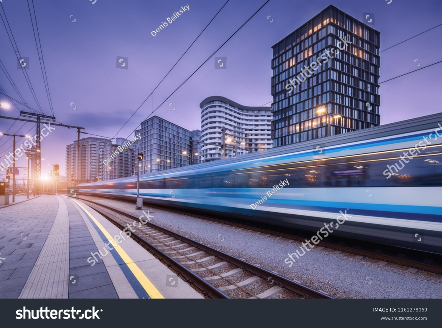 High speed train in motion on the railway station at sunset. Moving blue modern intercity passenger train, railway platform, buildings, city lights. Railroad in Vienna, Austria. Railway transportation #2161278069