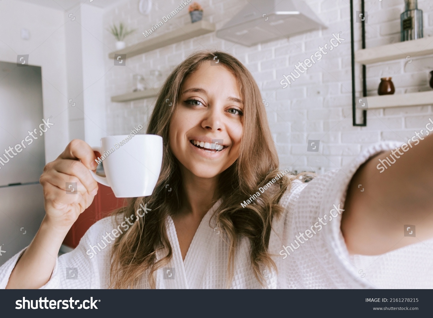 A young beautiful caucasian smiling blonde woman in a white robe with a white cup of coffee or tea takes a selfie in the morning on background of kitchen. Happy girl drinks hot beverage at home #2161278215