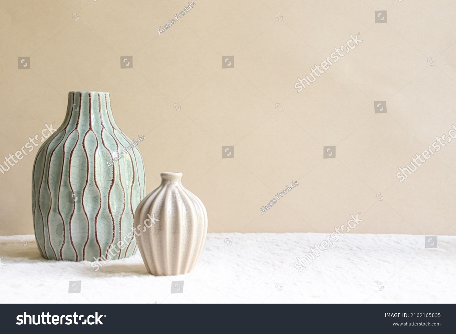 Duo of vases on a beige background. A medium sized sky blue vase with brown grooves and a small beige vase.  #2162165835