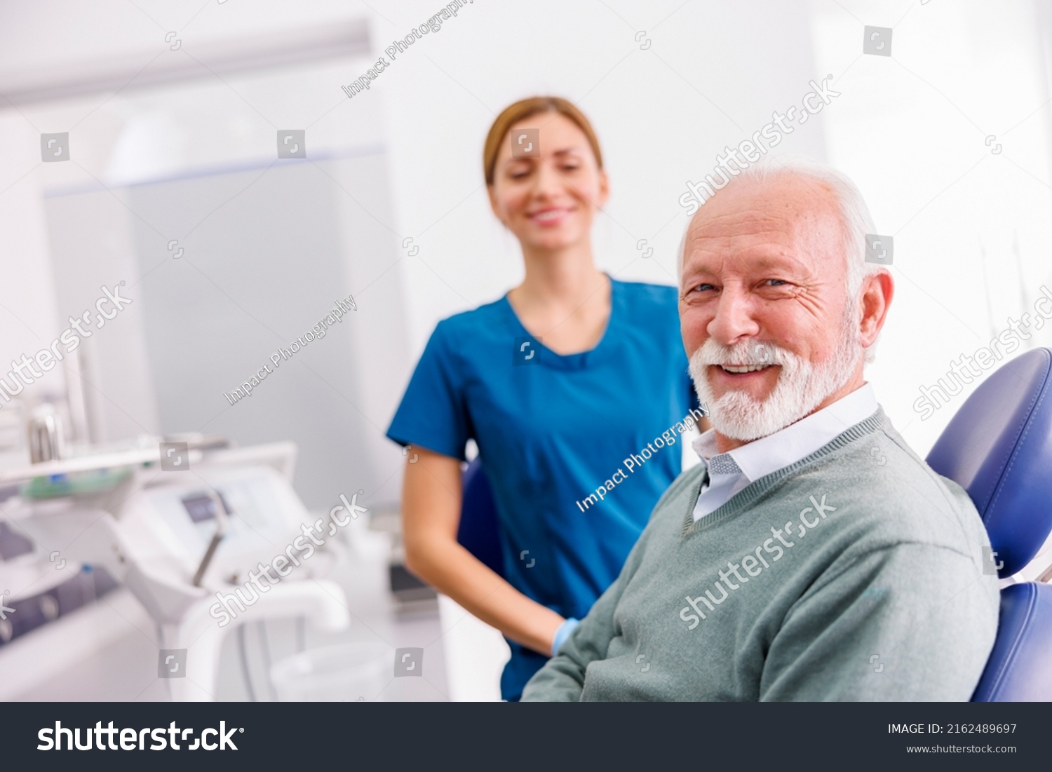 Portrait of satisfied senior male patient sitting at dental chair at dentist office smiling with doctor in the background #2162489697