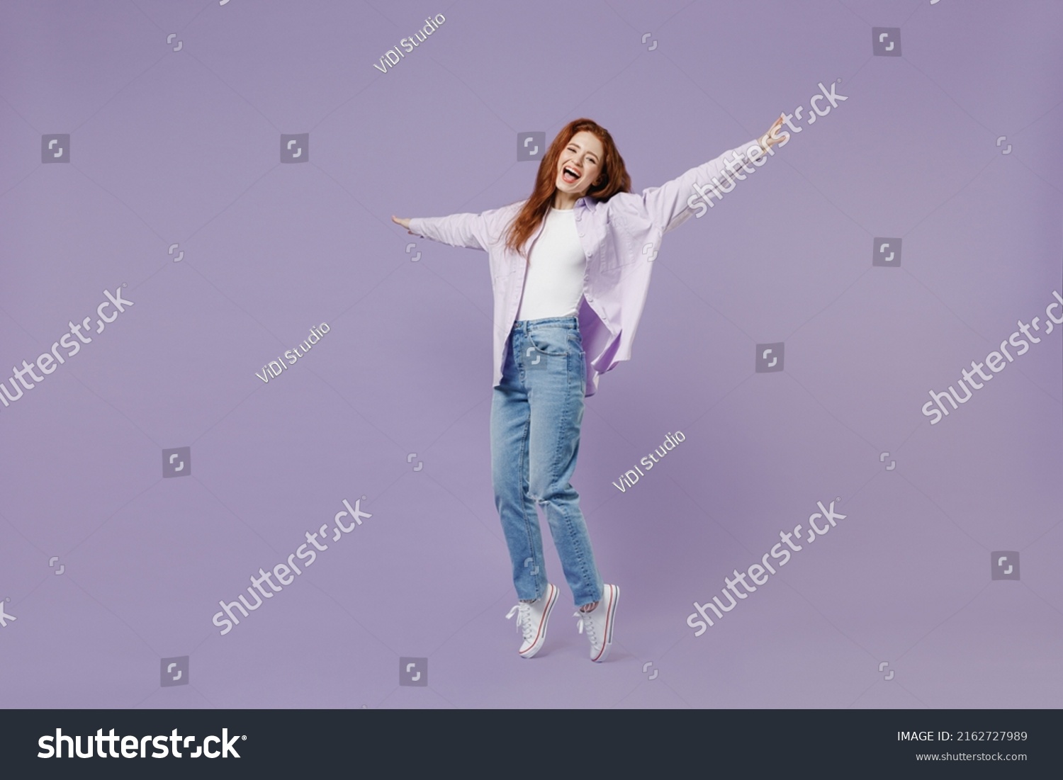 Full size body length young redhead curly woman 20s wear white T-shirt violet jacket stand on toes dance lean back have fun spread hands isolated on pastel purple color wall background studio portrait #2162727989