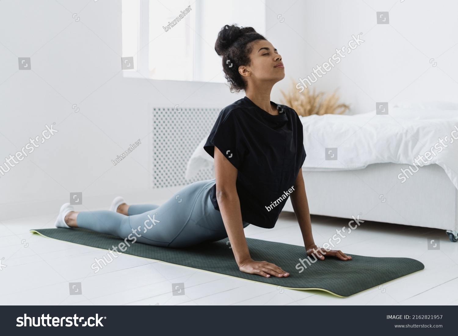 Side view of afro american female making breathing exercise, backbend and doing upward-facing dog pose or urdhva mukha svanasana. Woman at sport training at home, wearing in sportswear #2162821957