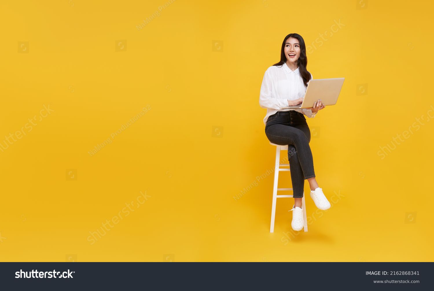 Young woman asian happy smiling. While her using laptop sitting on white chair and looking isolate on copy space yellow background. #2162868341