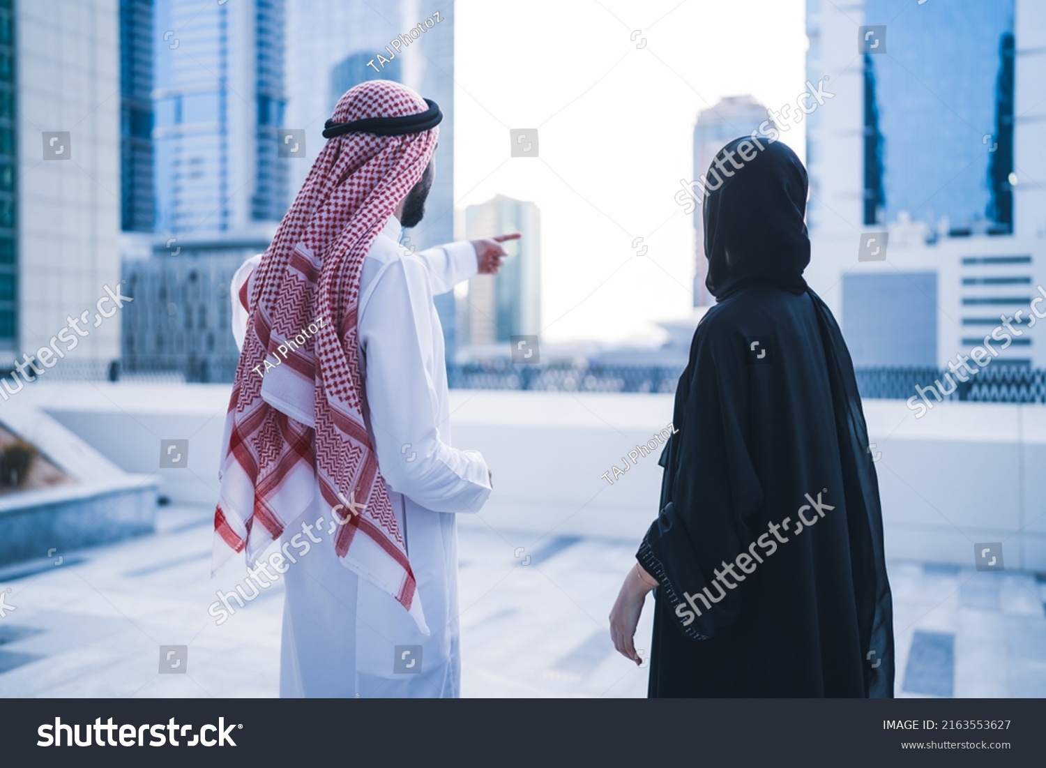 Back view of Arab family or business work colleagues pointing towards city skyline wearing traditional abaya and dress. Muslim Saudi or Emirati couple or team working together #2163553627