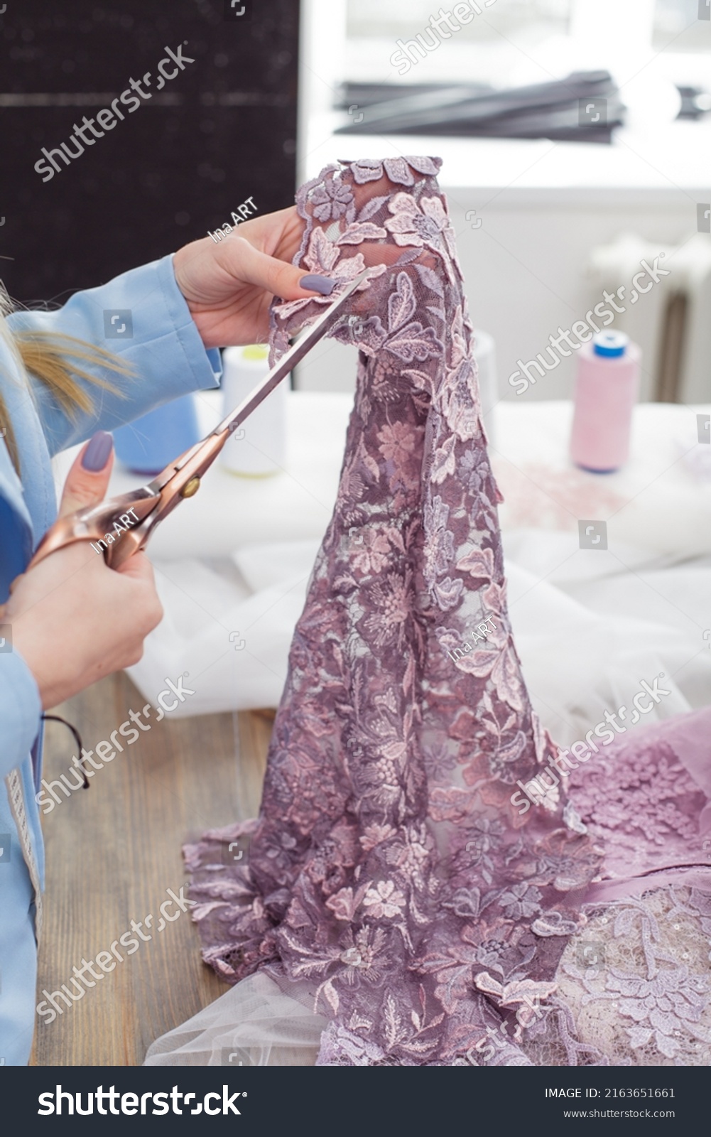 Tailor making a wedding dress with different lace and silk fabrics. Using scissors to cut purple lace. Working at her workshop. Cropped studio faceless portrait of Seamstress designer at workplace. #2163651661