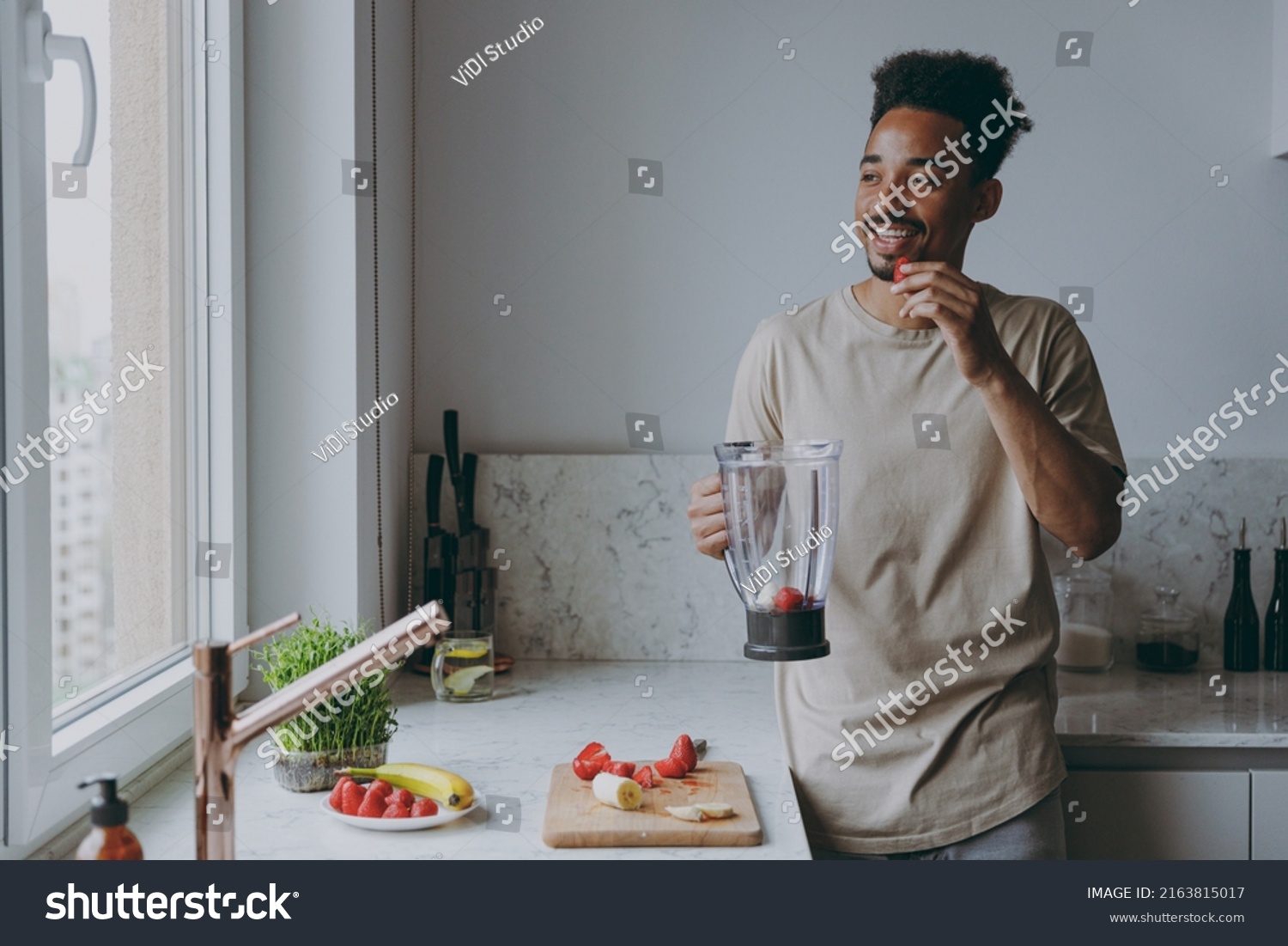 Young smiling african american man 20s in casual clothes prepare fruit cocktail eat strawberry using mixer blender cooking food in light indoor kitchen at home alone. Healthy diet lifestyle concept. #2163815017