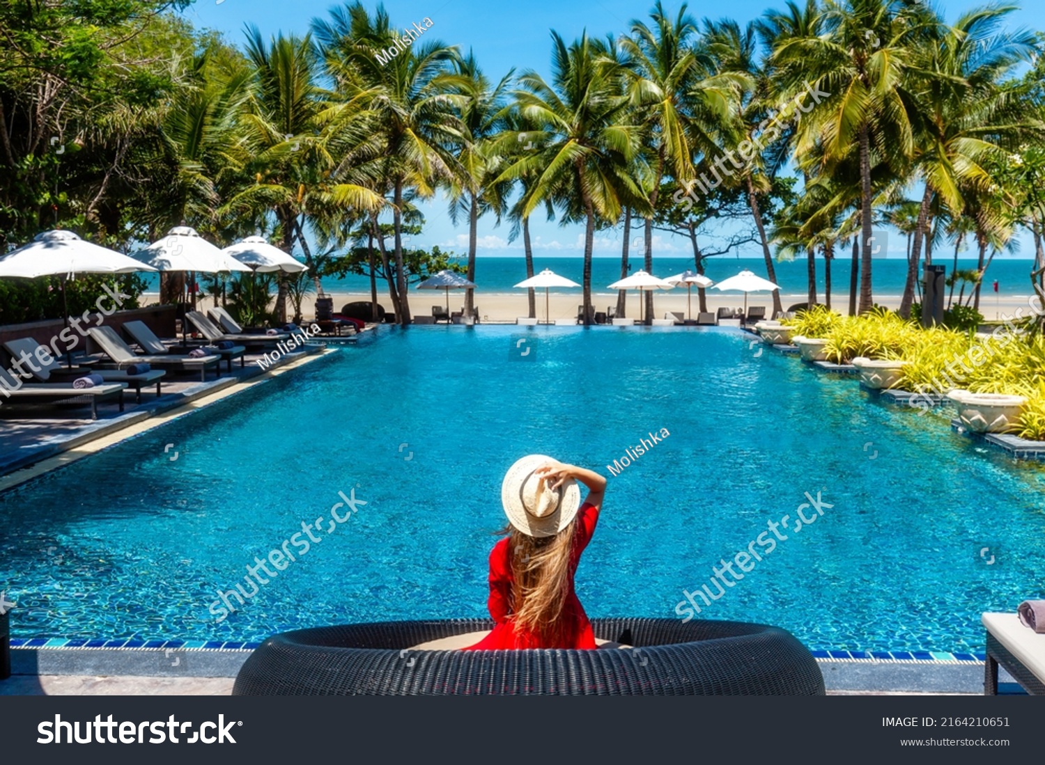 Luxury beach vacation in tropical beach hotel. Tourist woman in red dress relax near blue swimming pool in modern resort. Female traveler on sea vacation. #2164210651